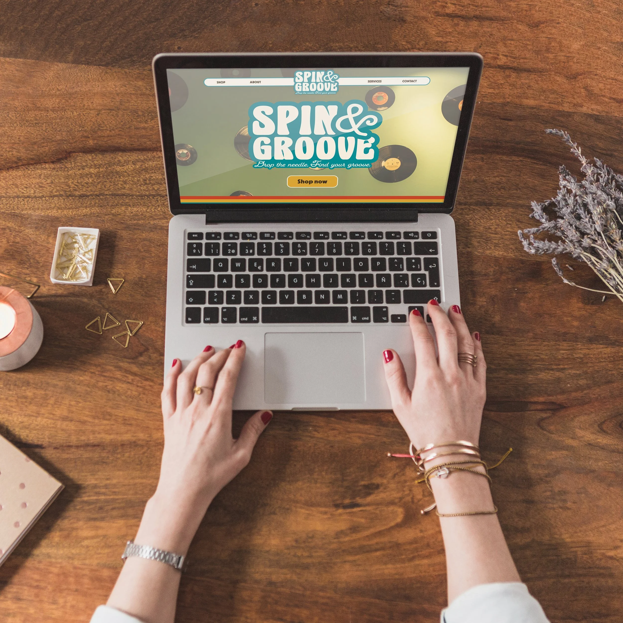 A person's hands with red nail polish using a silver laptop on a wooden desk, with a lavender bouquet, gold paper clips, a candle, and a closed notebook nearby. The laptop screen displays a website called 'Spin & Groove' with a colorful vinyl record theme.