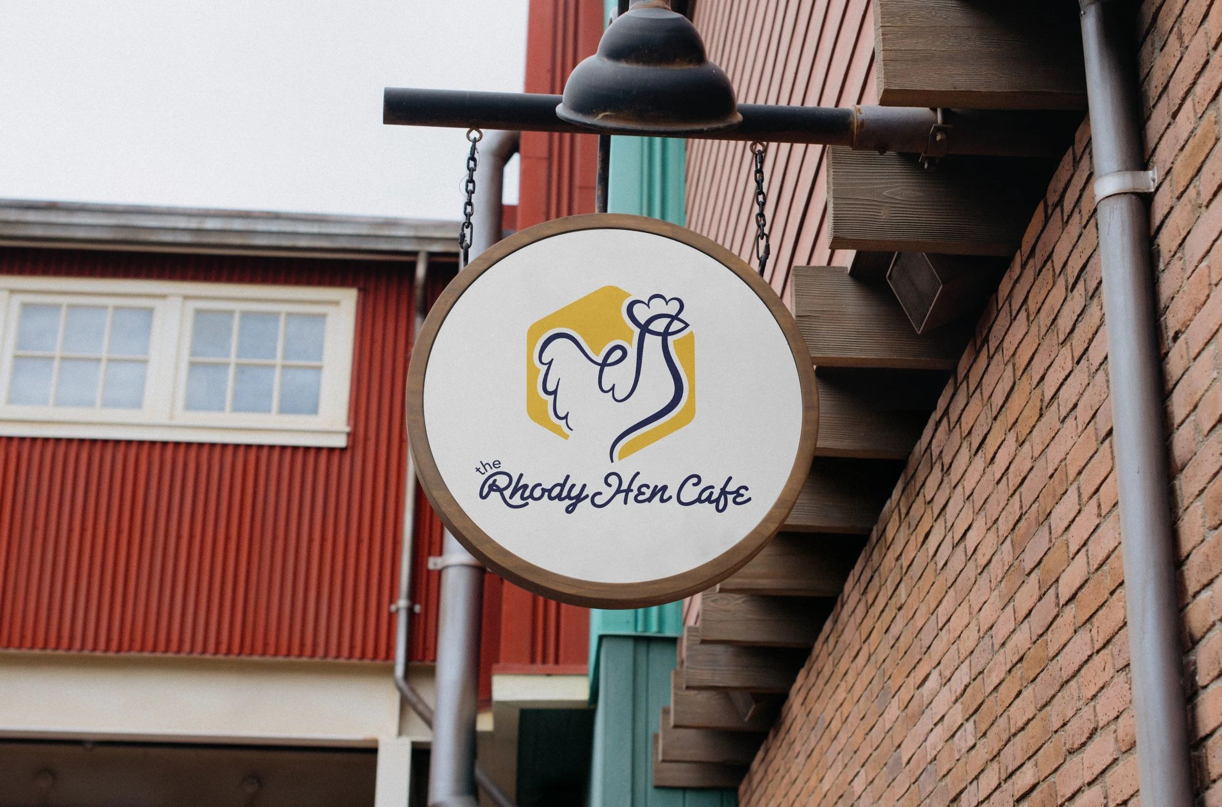Sign for Rhody Hen Cafe with a stylized hen logo on a white background, hanging outside a brick building.