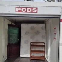 Storage unit with a sign labeled 'PODS', containing a mattress, a wooden shelf, and a green object.