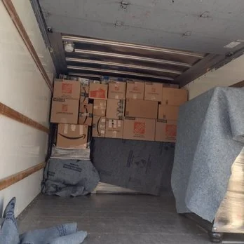 Inside of a moving truck with stacked cardboard boxes and a gray blanket or tarp on the floor.