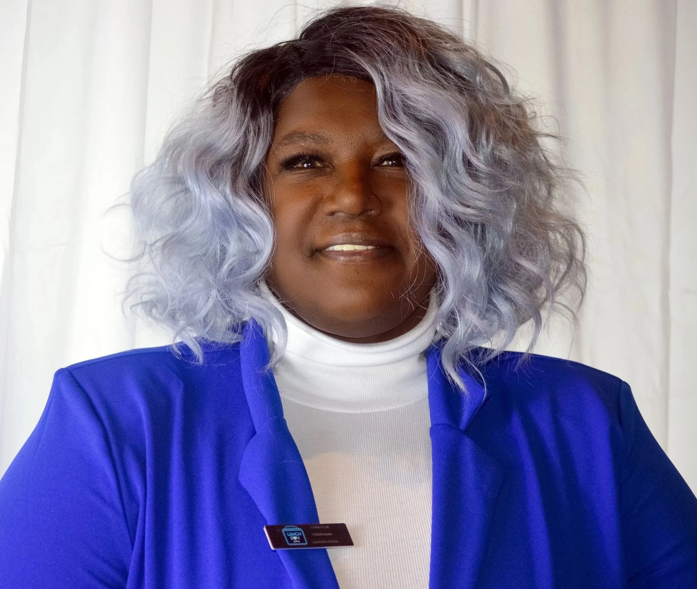 A woman with a warm smile and wavy, silver-colored hair, wearing a blue blazer and a white turtleneck, standing in front of a white curtain.