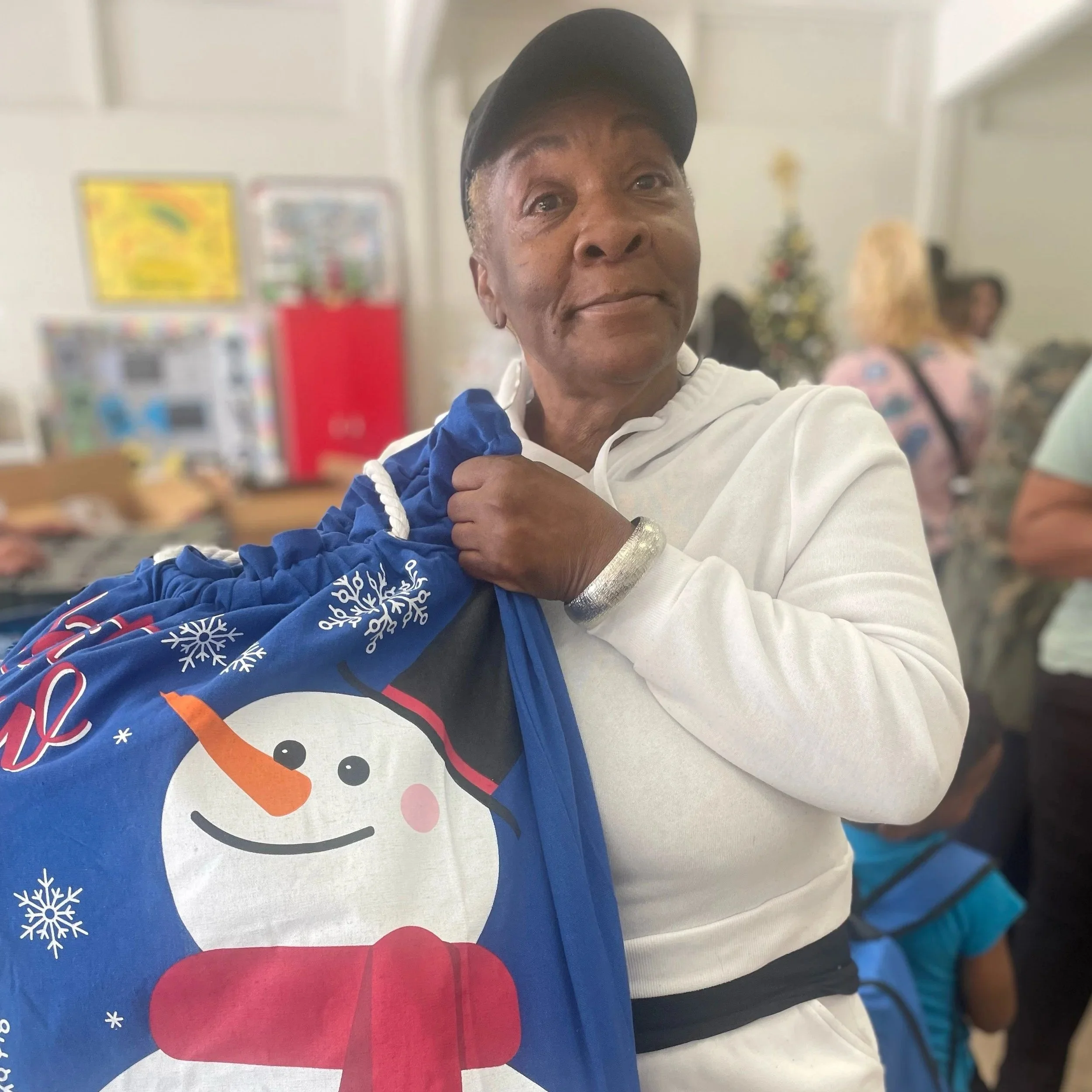 An elderly woman holding a blue Christmas-themed gift bags with a snowman design inside a busy indoor space, with other people in the background.