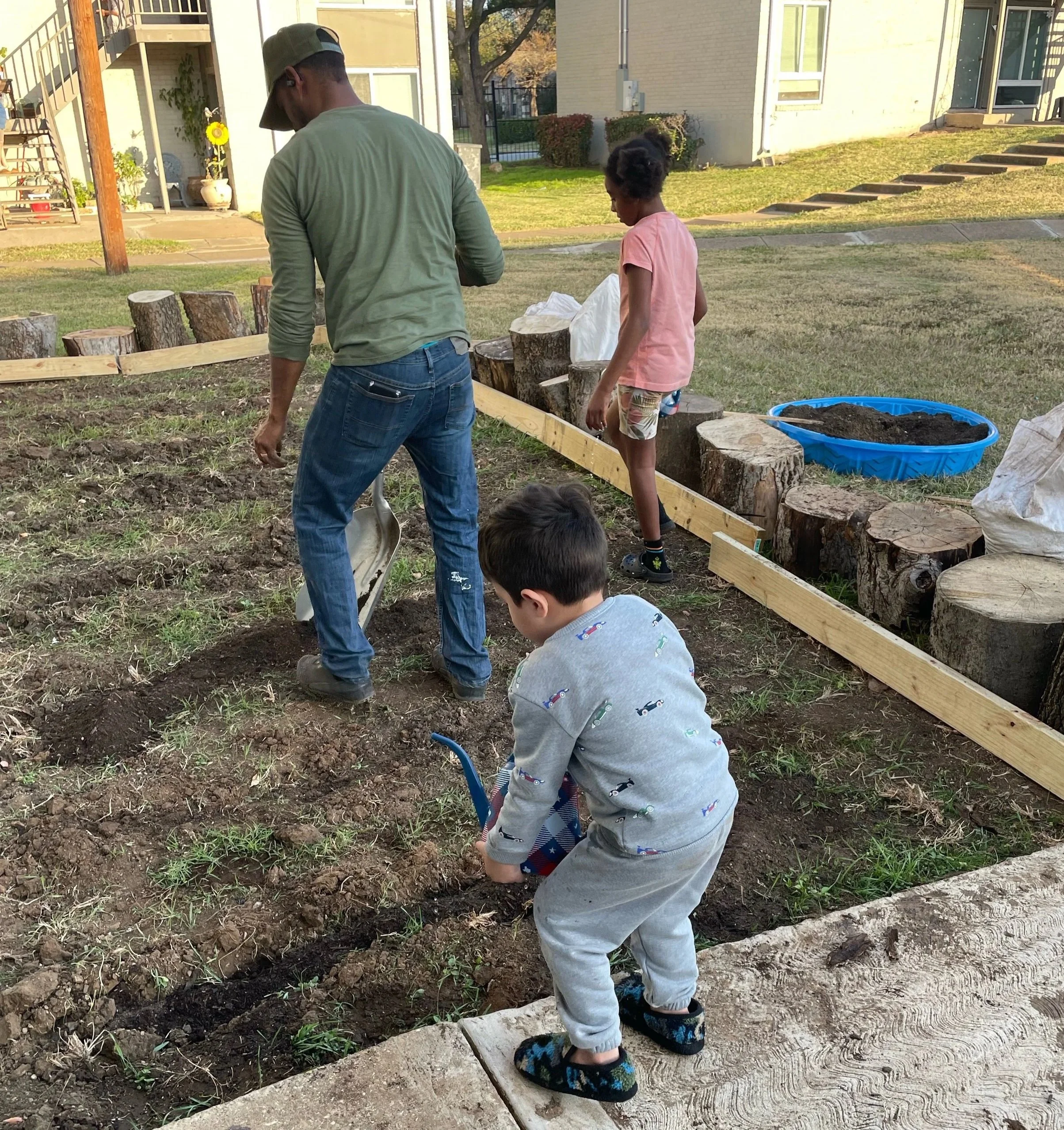 A man and three children working together in a garden, planting or digging in the soil, with tree stumps and a blue water container nearby.