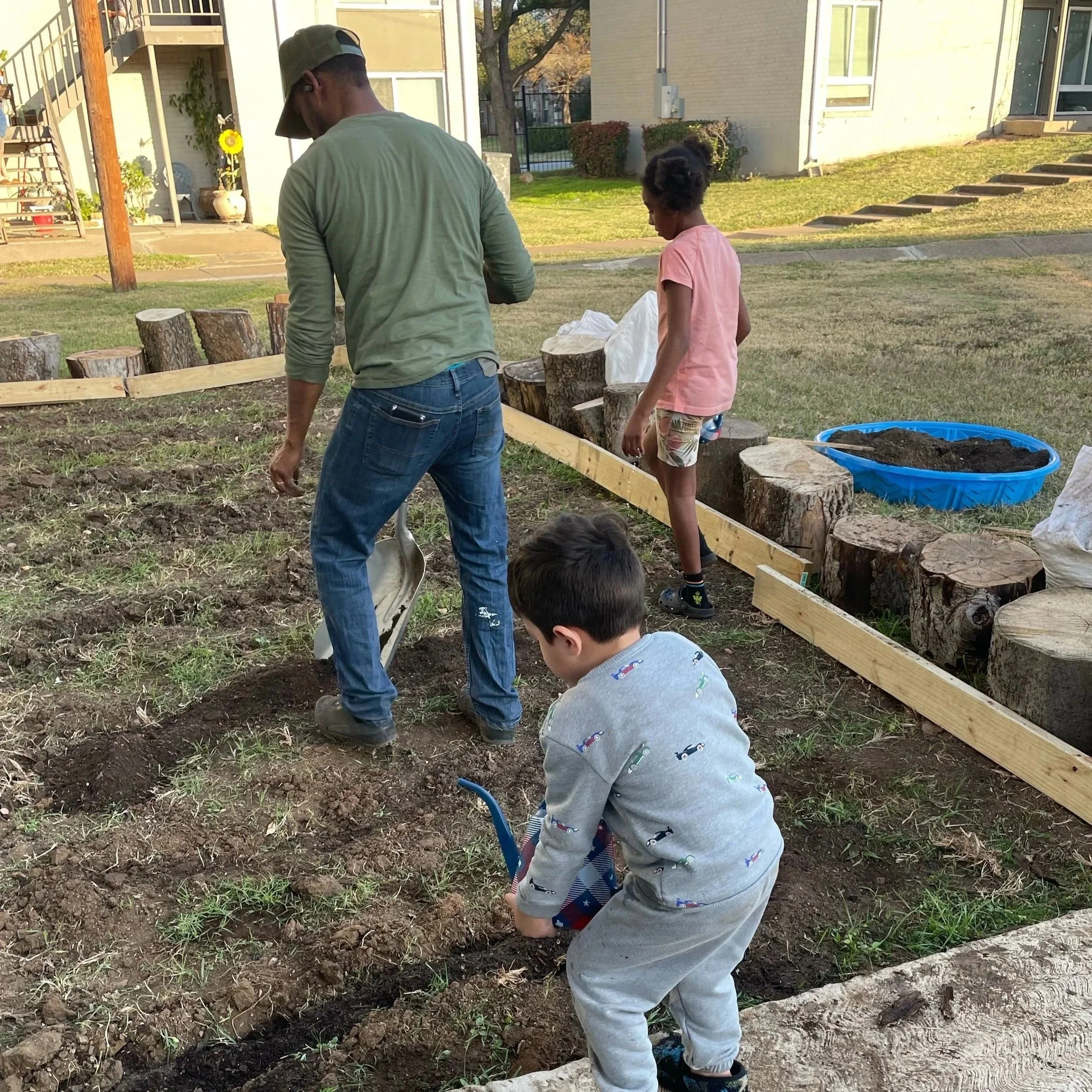 A man and three children working together in a garden, planting or digging in the soil, with tree stumps and a blue water container nearby.