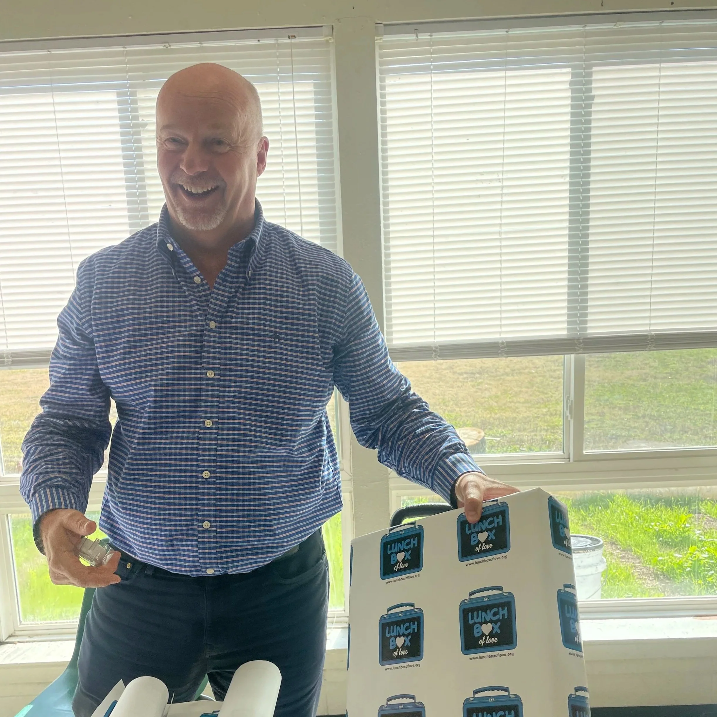 Man smiling and holding a lunch box with a matching sign that says 'Lunch Box of Love' in a bright room with large windows.