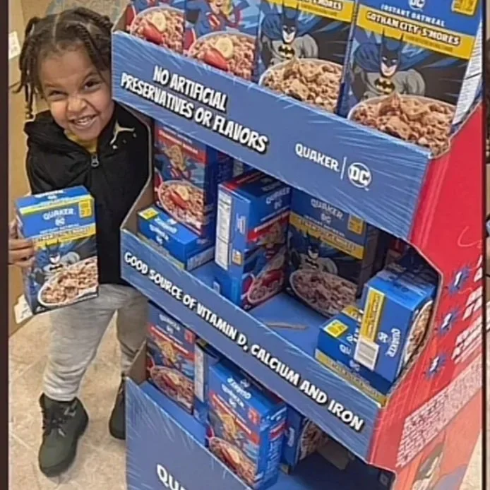 Two young girls smiling and holding Quaker oatmeal cereal boxes next to a retail display of Quaker oatmeal products featuring Batman branding from DC Comics.