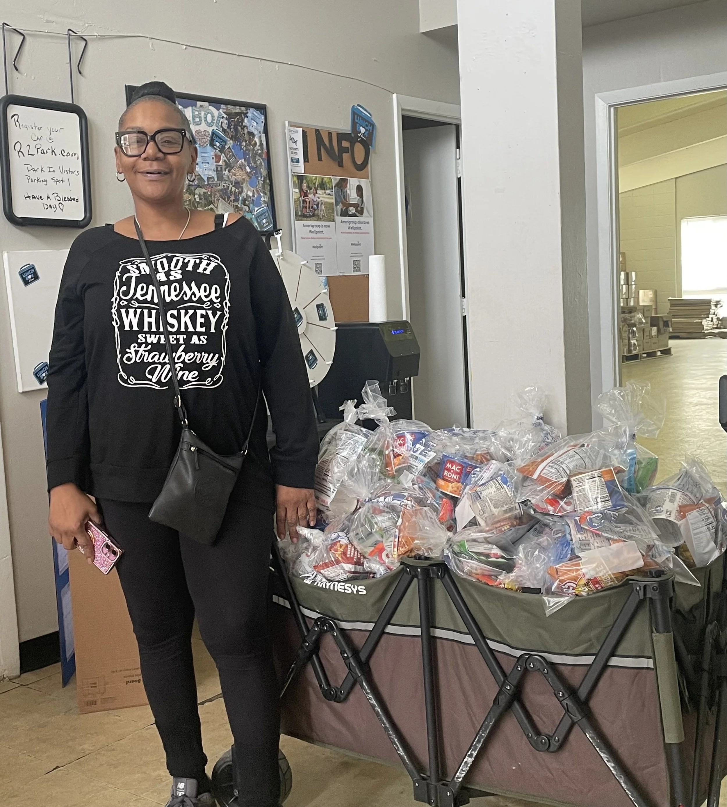 Smiling woman standing next to a cart filled with plastic bags of canned goods inside a room with a bulletin board and a doorway leading to another room.