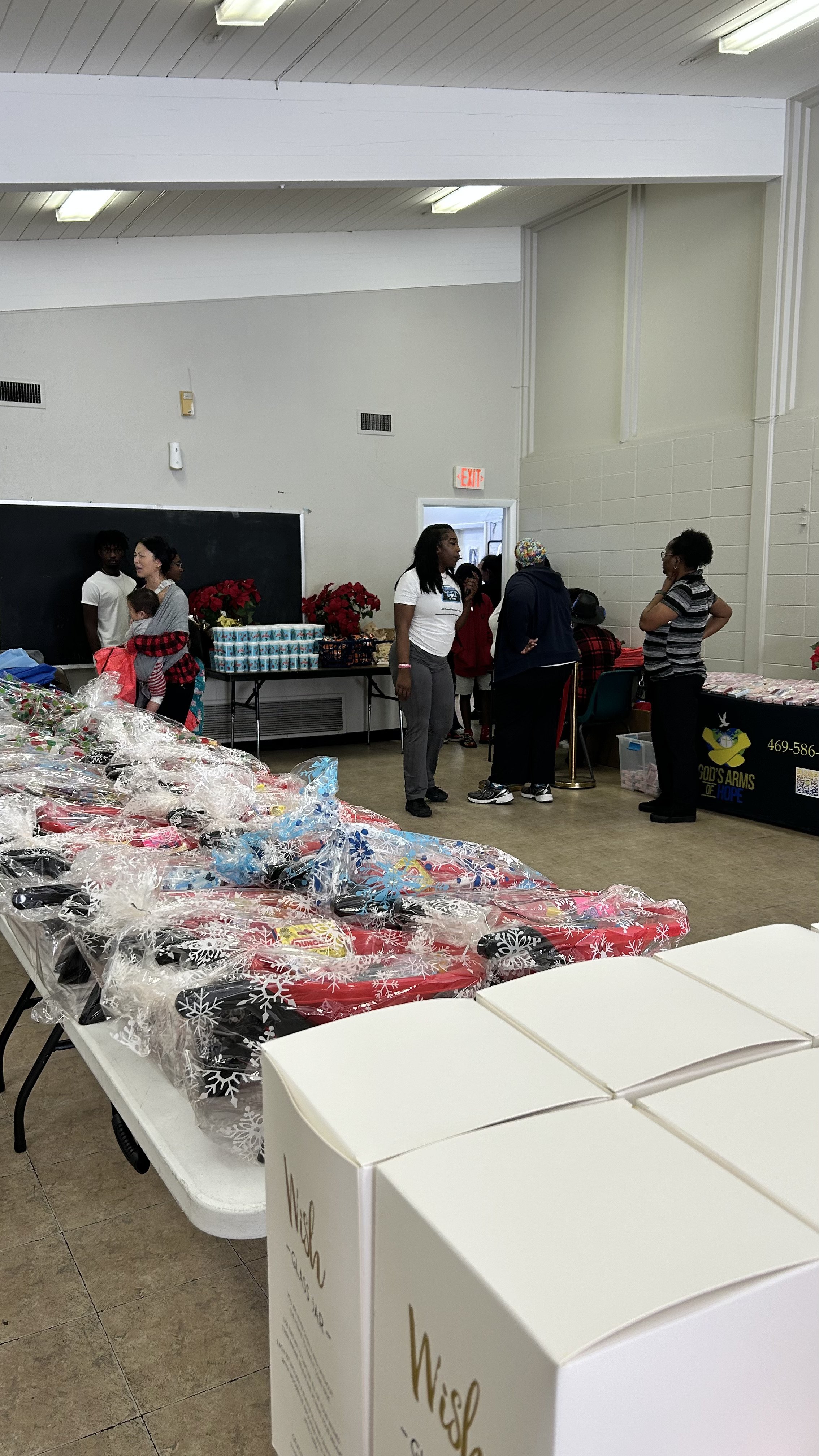 Indoor community event with tables displaying wrapped gifts, Christmas poinsettias, and various items, with people engaging in conversations.