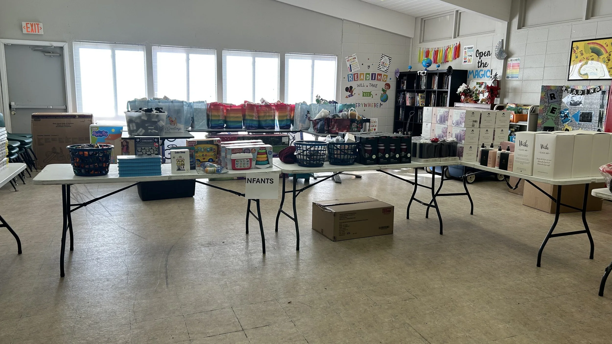 Tables with toys, books, and household items set up for a sale or donation event in a community center room with windows, chairs, and colorful decorations.