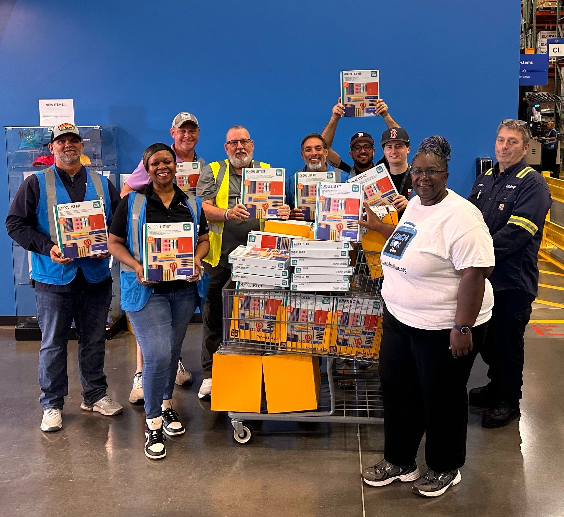 A group of ten people standing in a store, smiling and holding school supply boxes. They are gathered around a shopping cart filled with more school supplies, with a bright blue wall in the background.