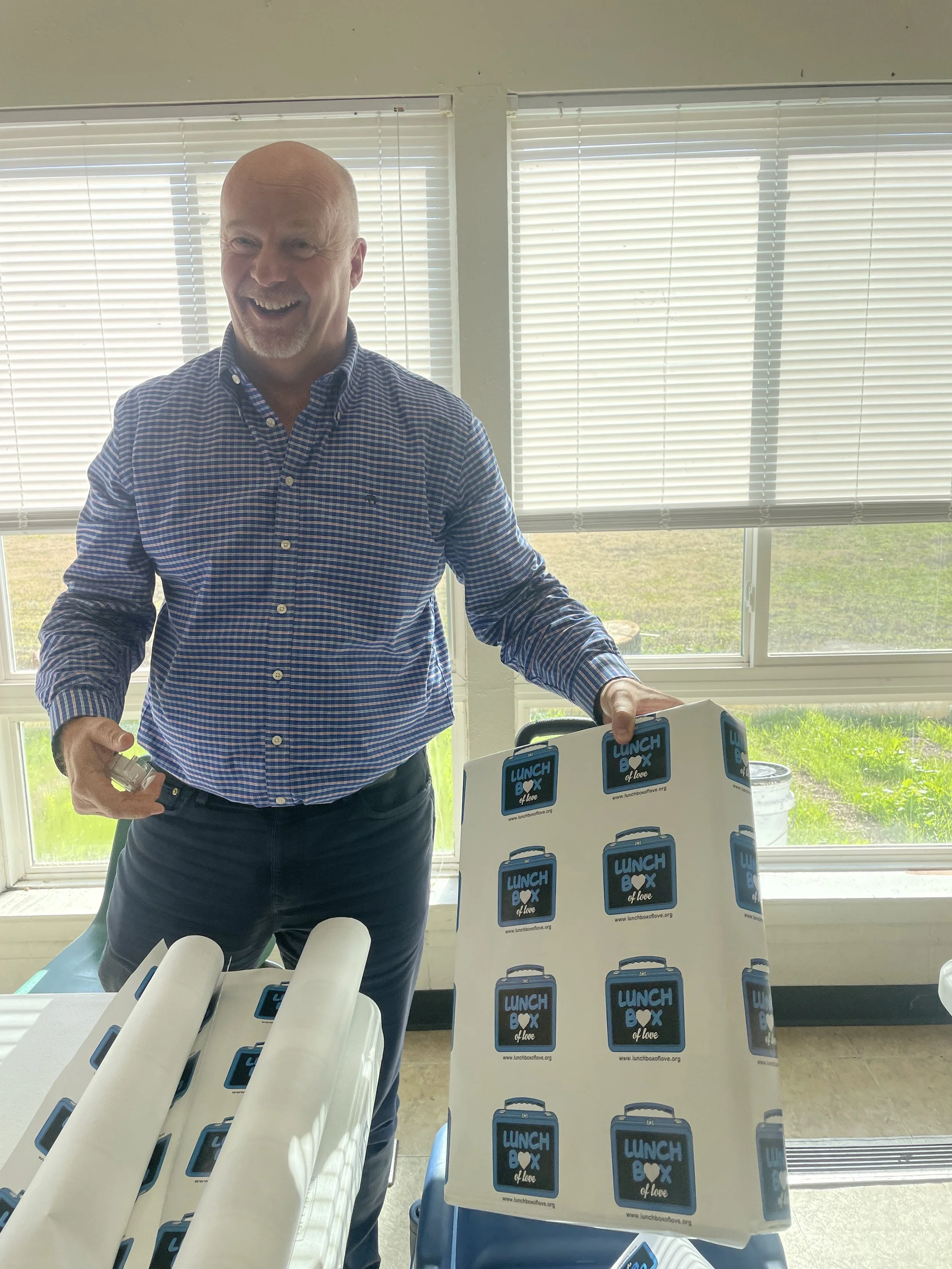 Man smiling and holding a lunch box with a matching sign that says 'Lunch Box of Love' in a bright room with large windows.