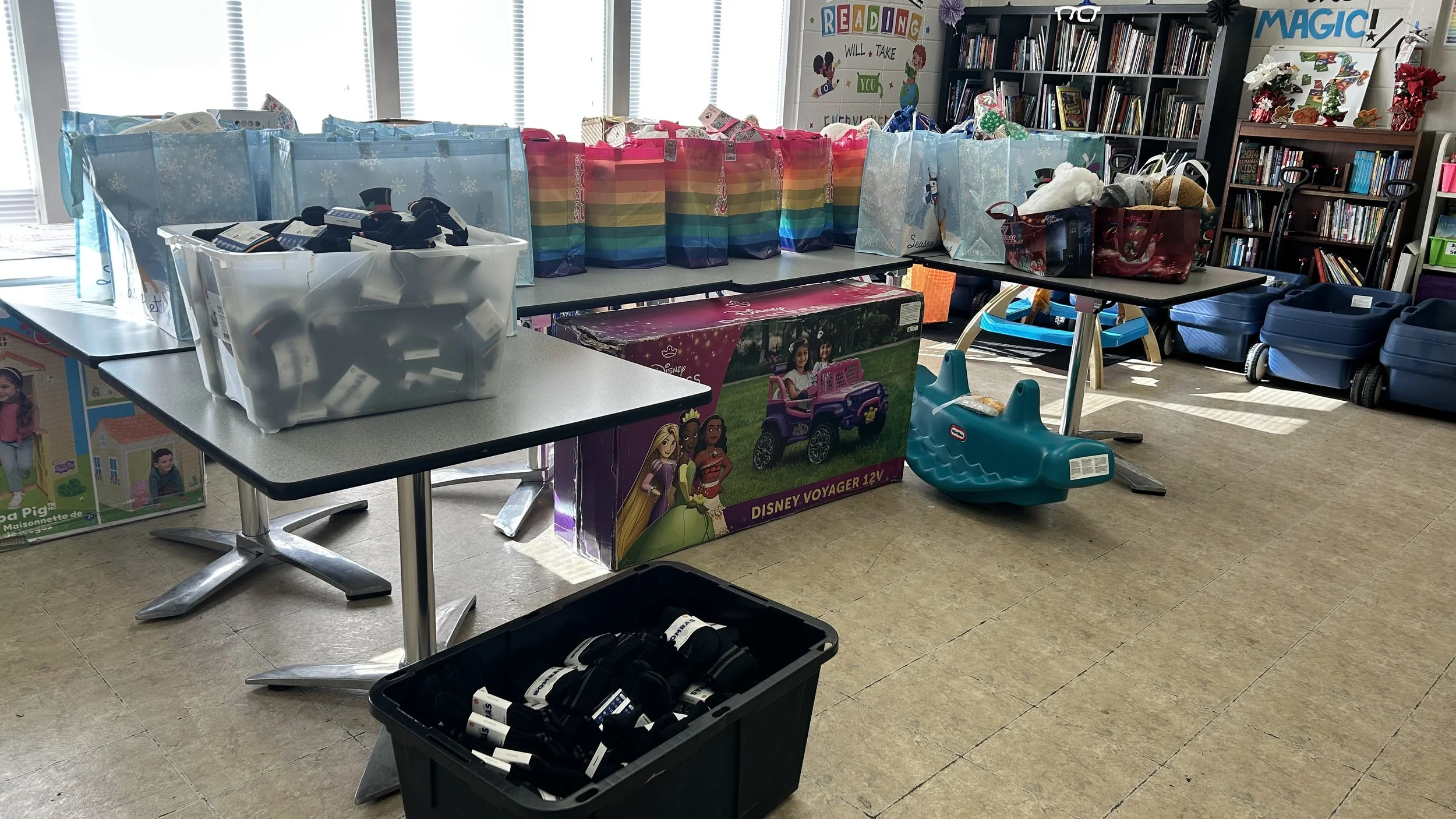 Tables filled with gift bags, toy boxes, and children's toys in a room with bookshelves, a colorful sign that says "READING will take you everywhere," and windows with blinds.