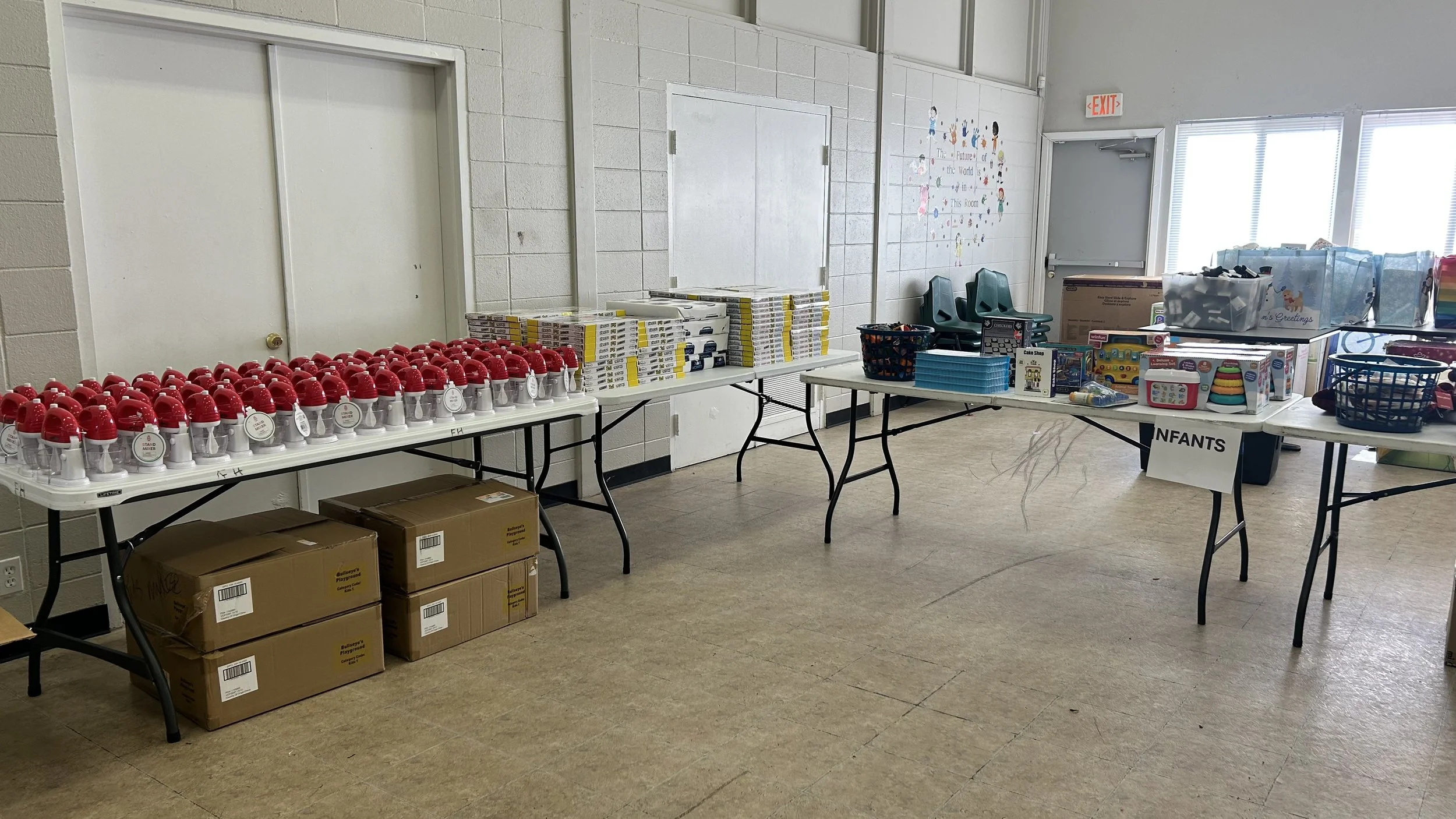 Tables set up in a room with various donated items, including red and white toys, boxes of supplies, baskets with children's toys, and a sign labeled 'INFANTS'.