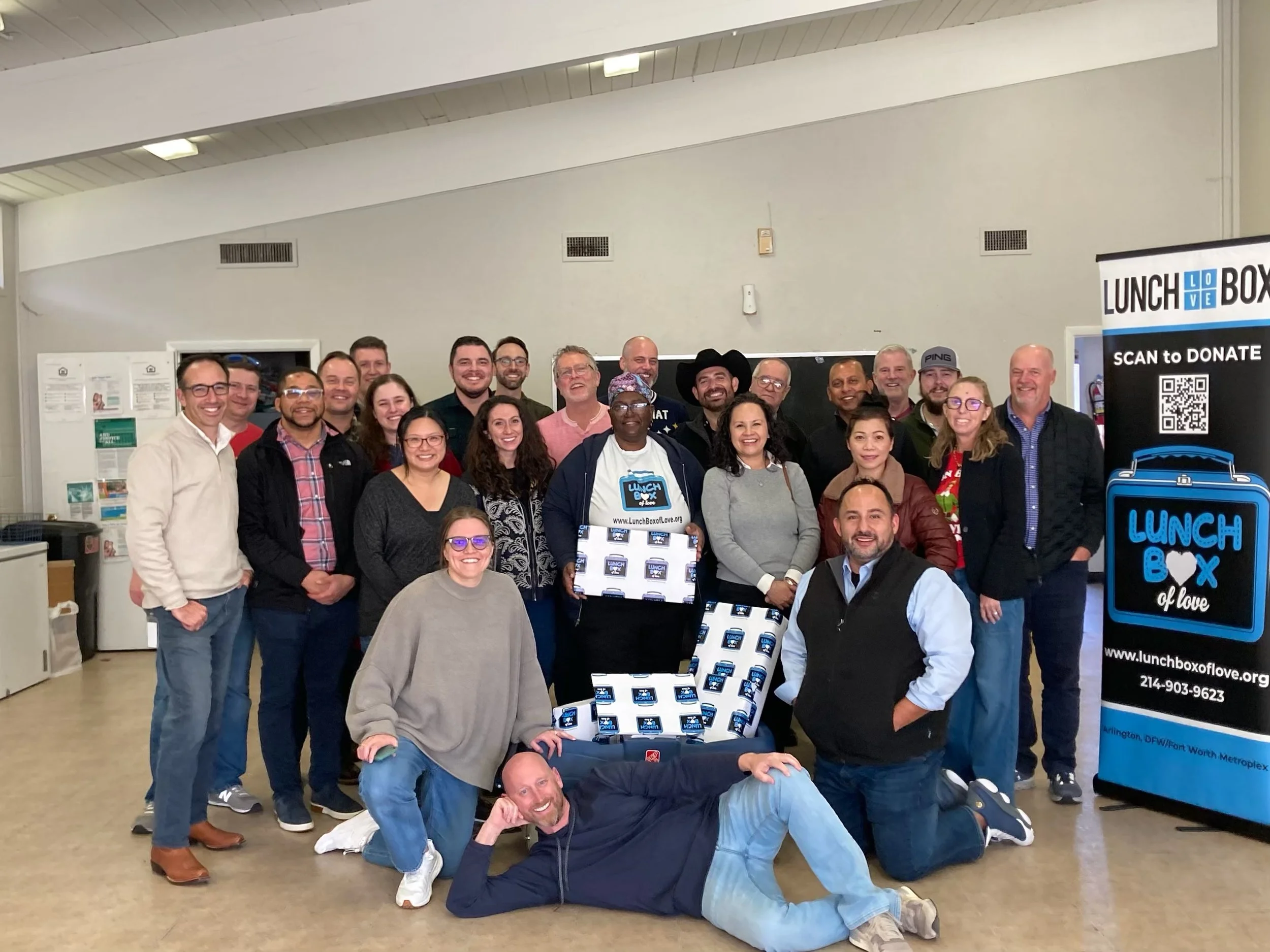 Group of diverse people gathered indoors for a photo, with a man in center holding a box and a sign with a website, next to a banner reading 'Lunch Box of Love' and 'Scan to Donate'.