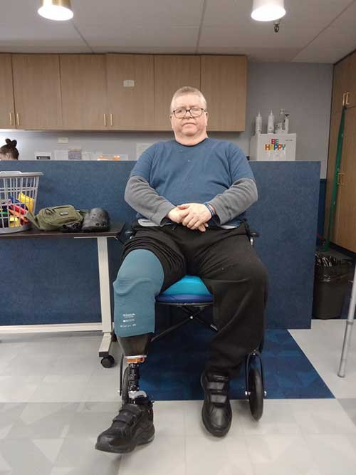 A man with a prosthetic leg sitting on a chair in an indoor setting, with a blue and gray background.