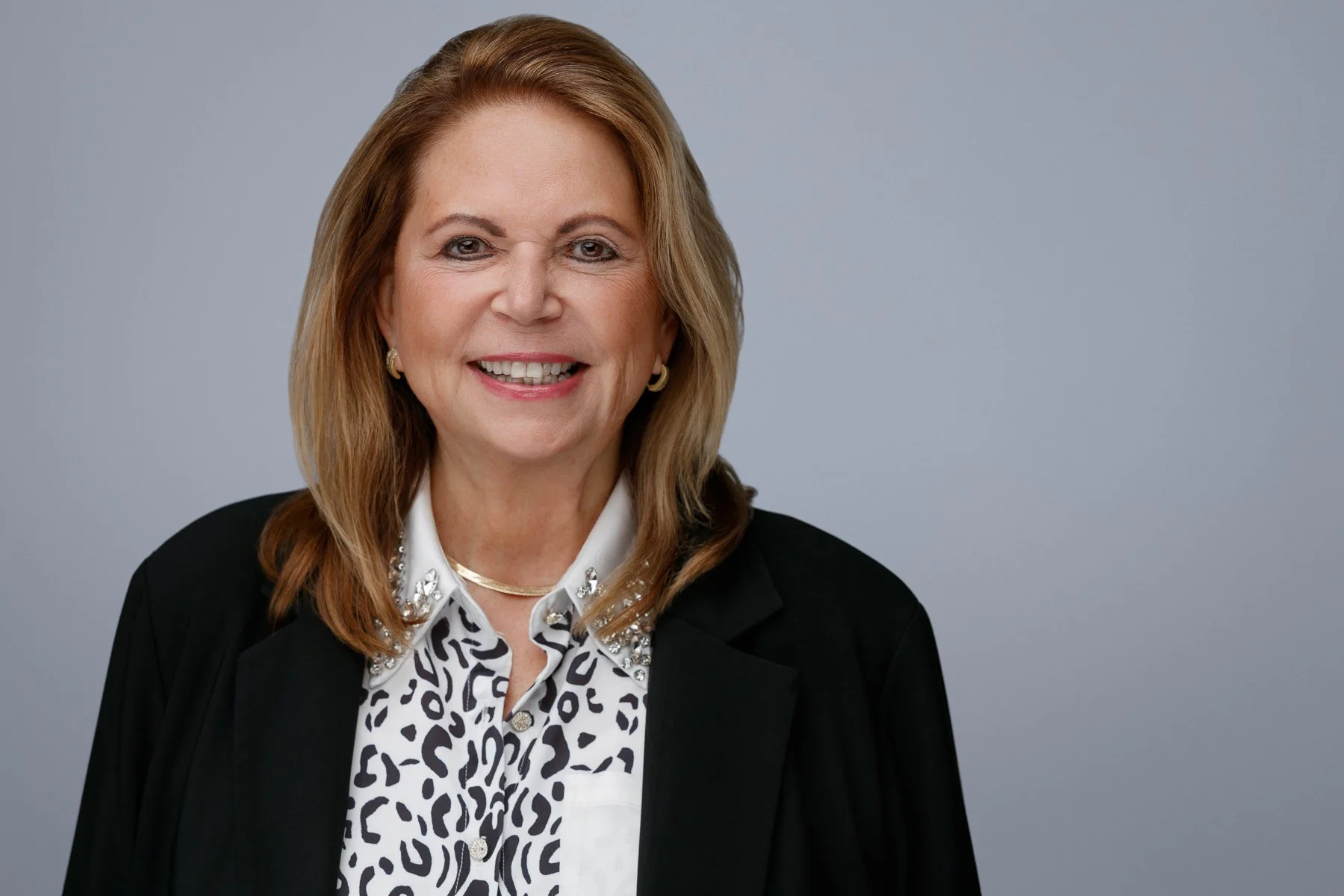 A middle-aged woman with shoulder-length, light brown hair smiling, wearing a white blouse with a black animal print pattern, a black blazer with embellishments on the lapel, and gold jewelry, against a plain light gray background.