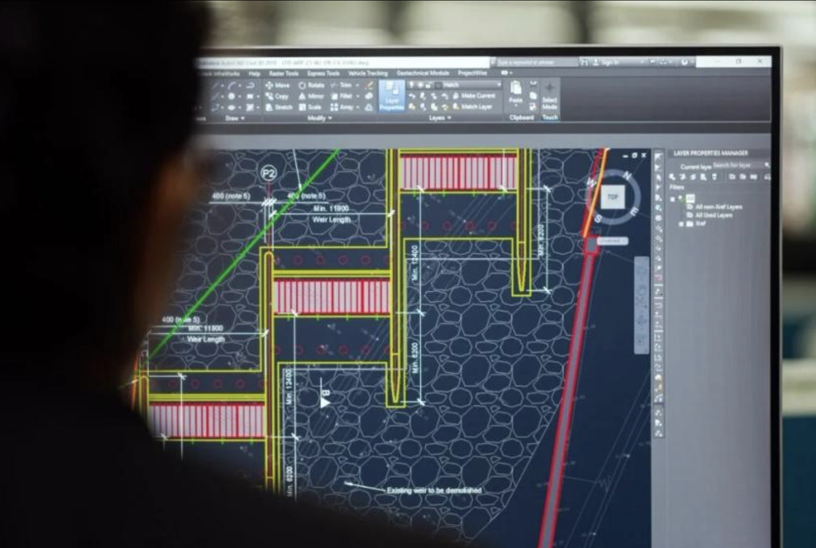 A person viewing a computer screen displaying a detailed architectural or engineering drawing of a landscape or construction plan. The diagram includes measurements, color-coded sections, and labels related to site layout.