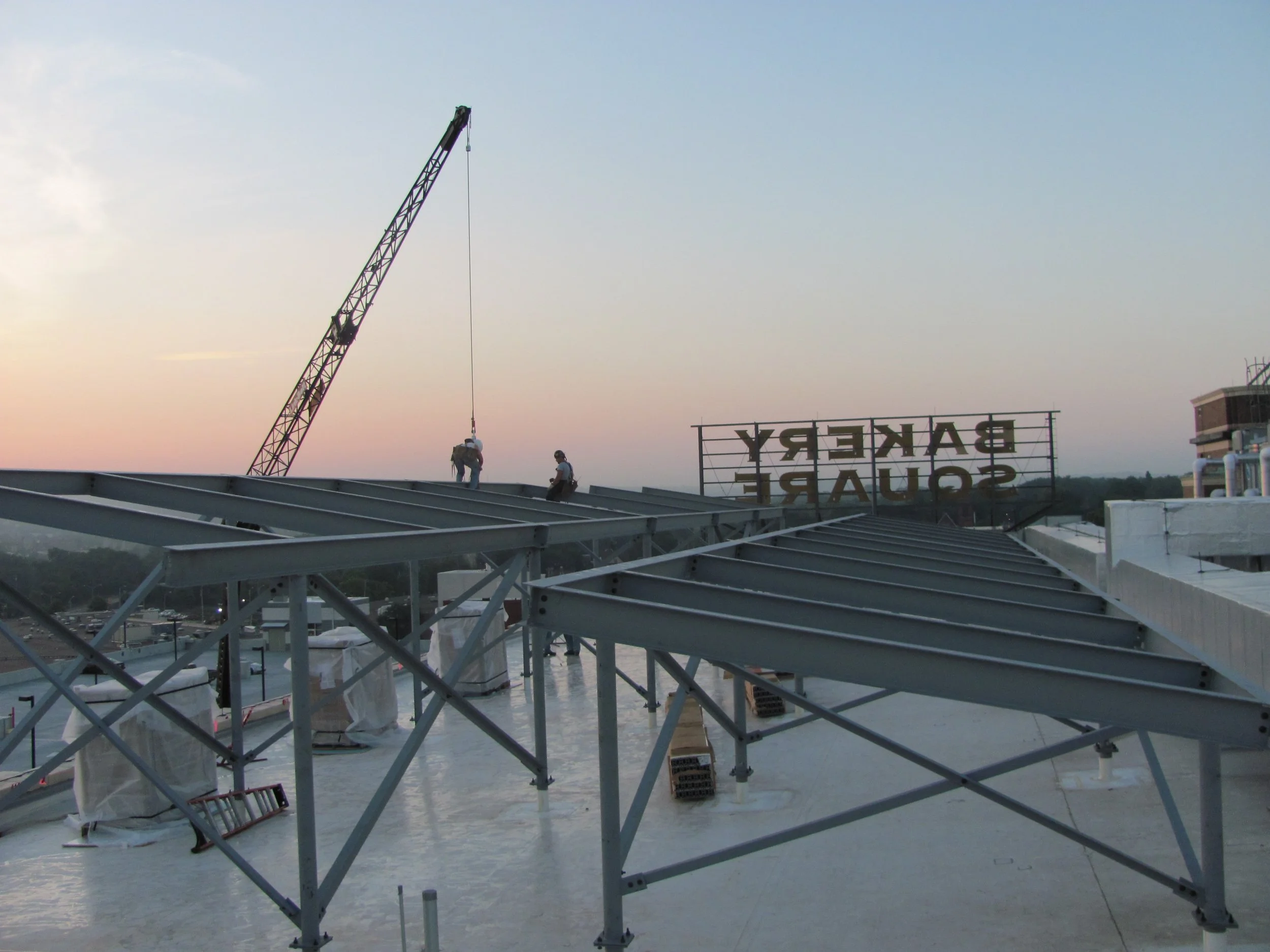Construction workers on a rooftop installing or maintaining a large sign banner that reads "BAKERY SQUARE" at sunset, with a crane in the background.