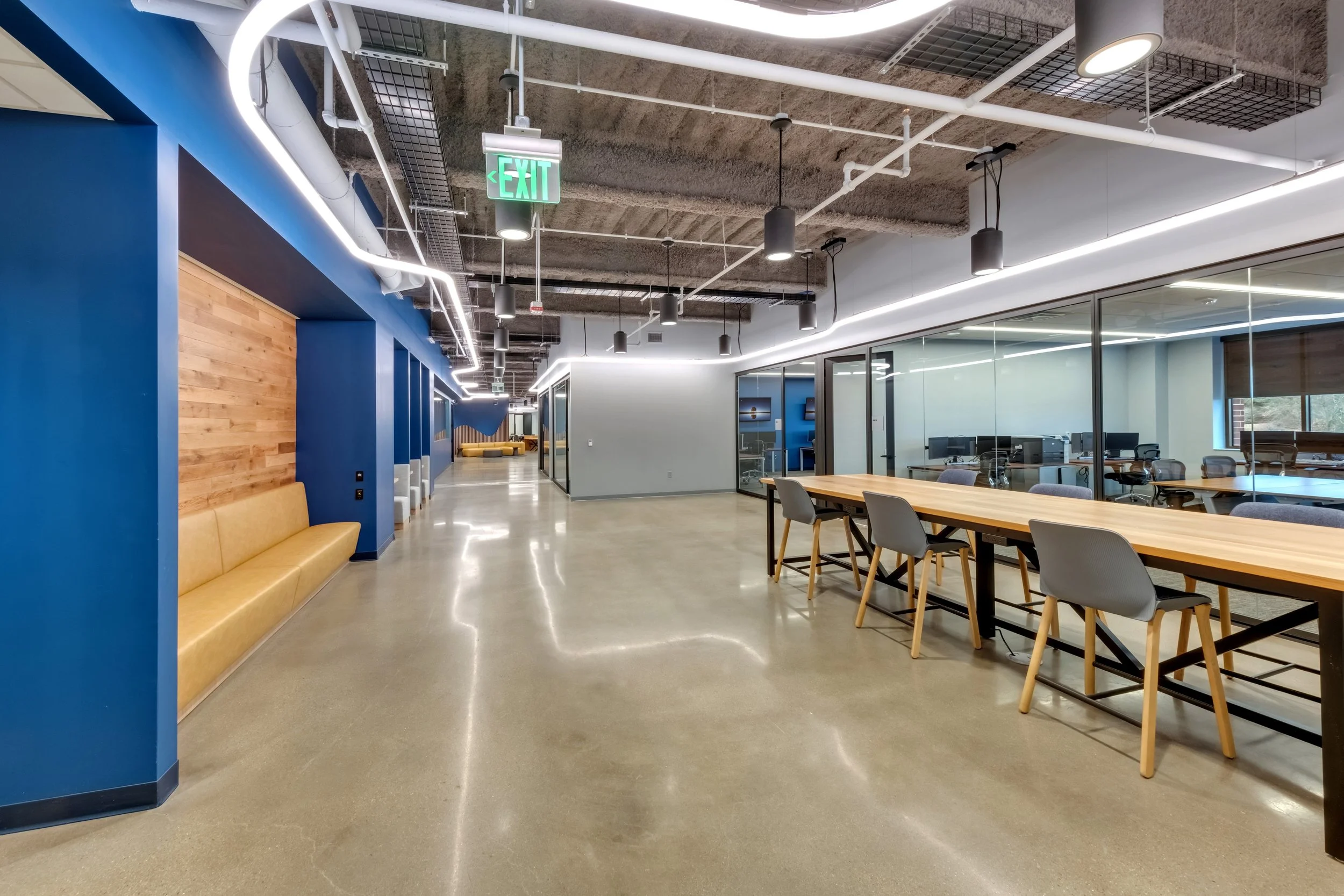 Modern office interior with communal tables, seating area, glass enclosed meeting rooms, polished concrete floors, exposed ceiling with industrial lighting, and a yellow bench against a blue wall.