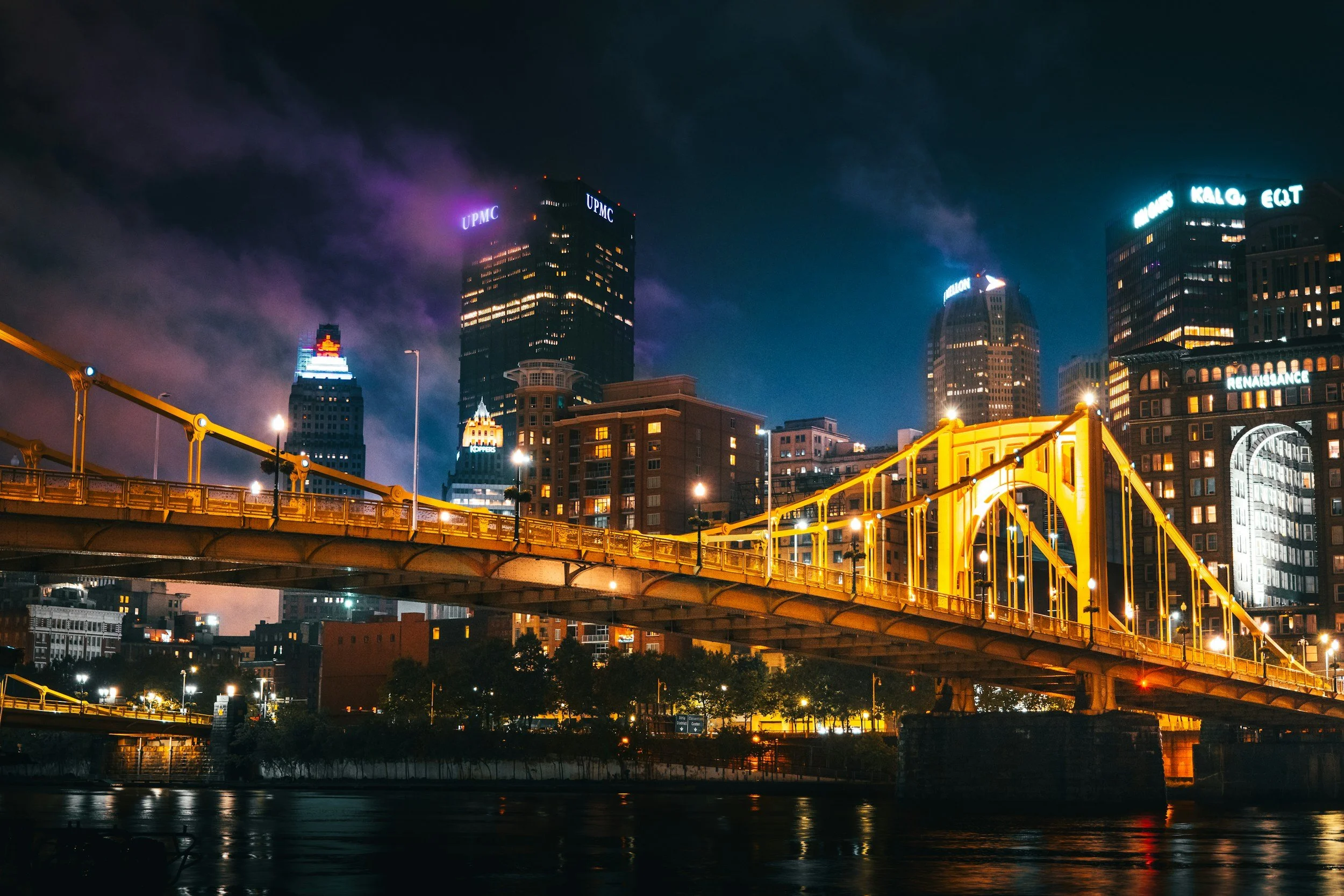 Night view of a city skyline with illuminated buildings and a yellow bridge over a river.
