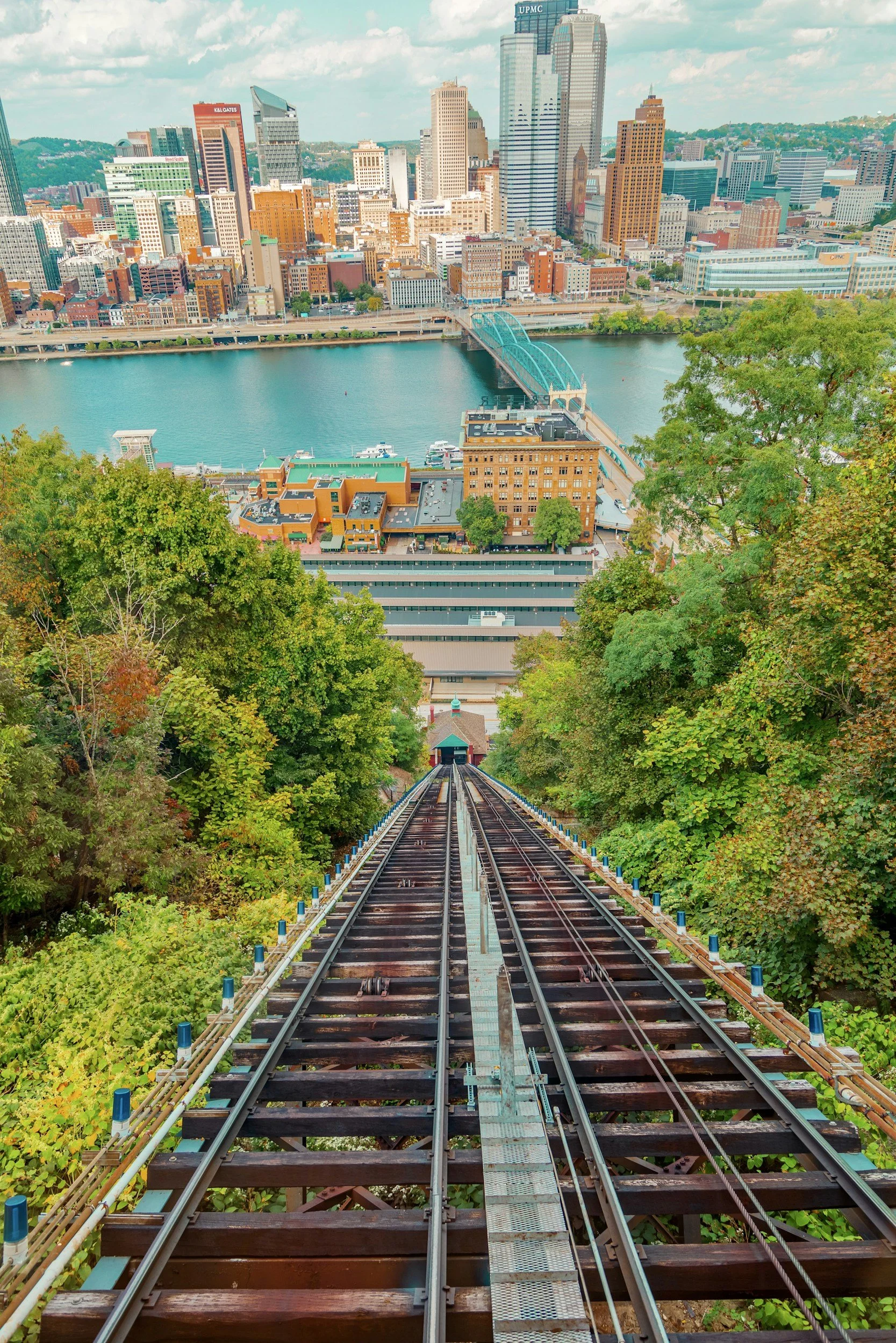View from a steep sky tram cable car track descending through green trees towards downtown Pittsburgh, Pennsylvania, with the city skyline and river in the background.