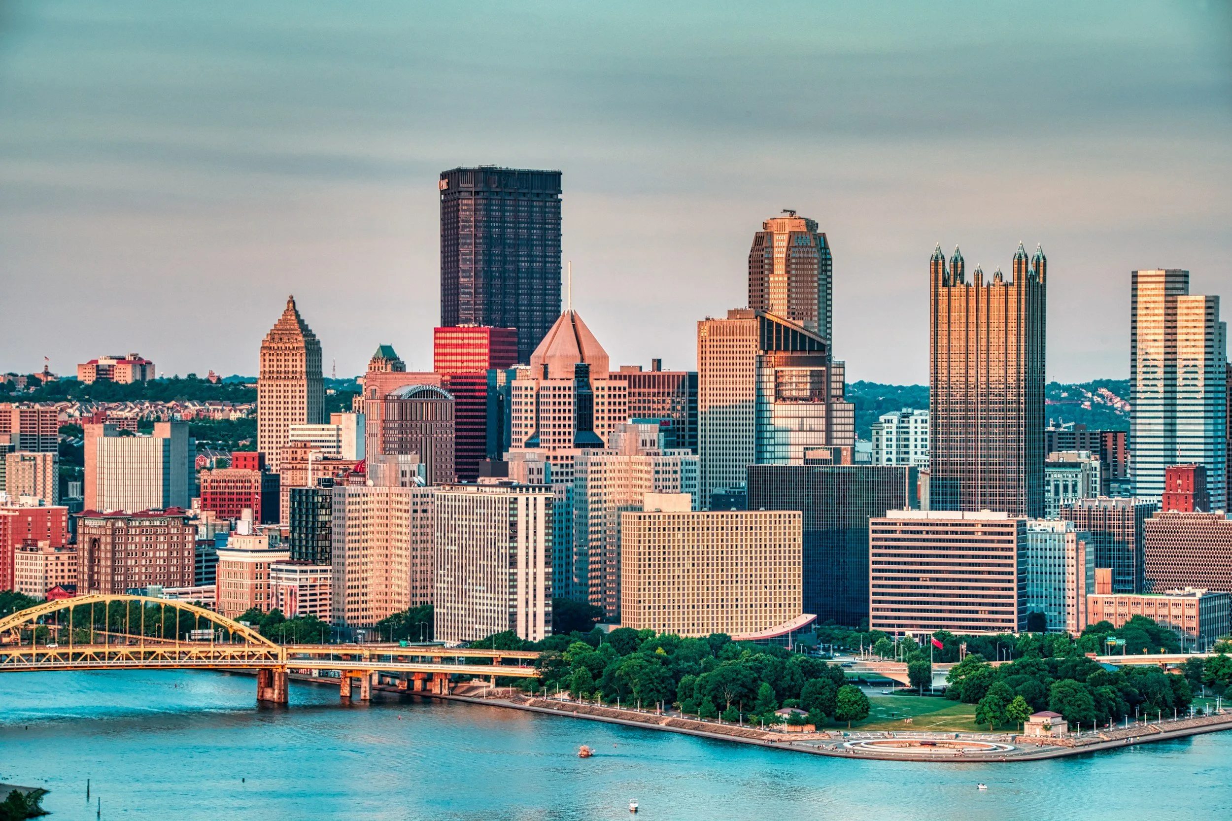 City skyline with tall skyscrapers along a river, featuring a yellow bridge and green park area in the foreground.