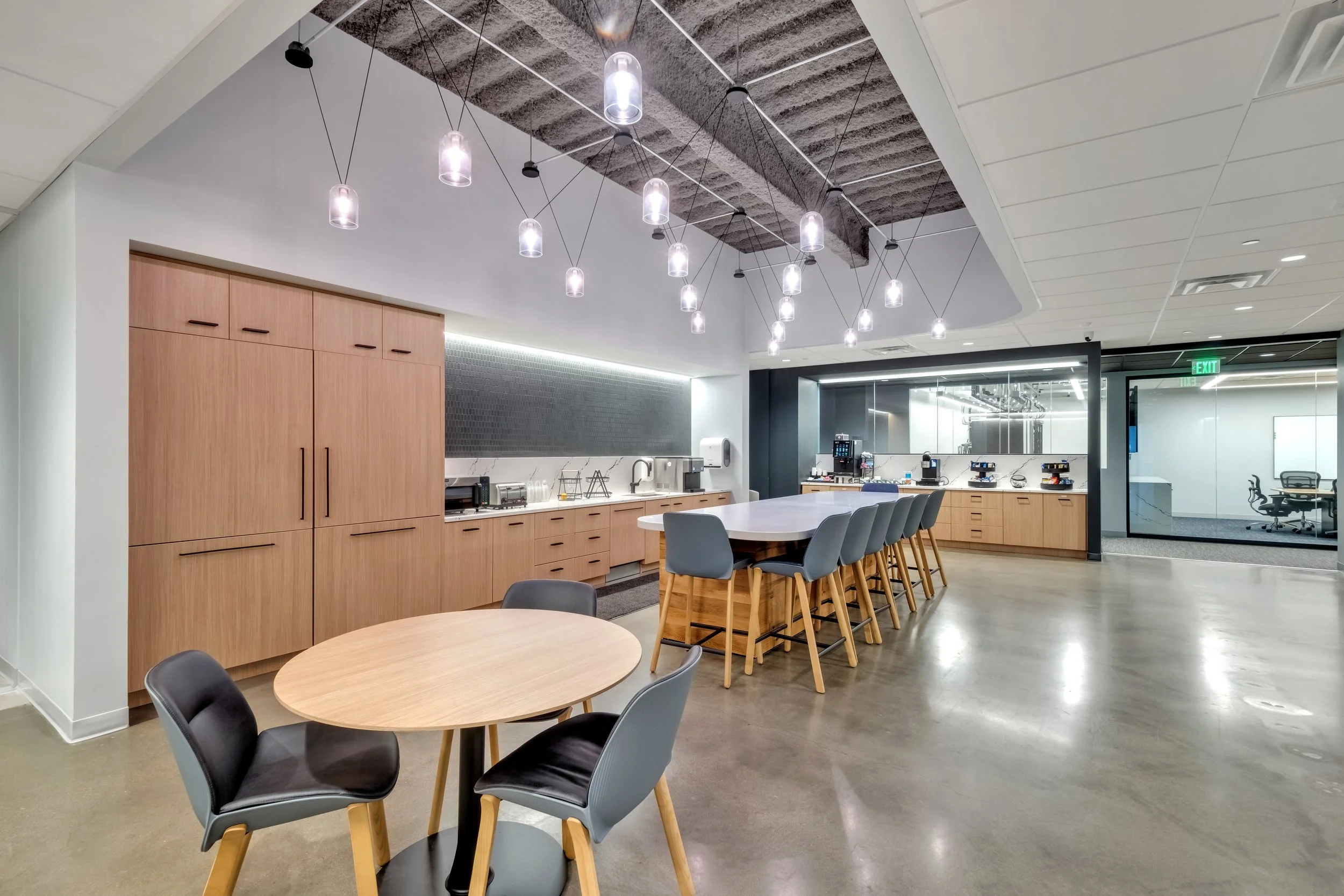 Modern office kitchen with wooden cabinets, a white island counter with gray chairs, and a coffee area with coffee machines. There are minimalist pendant lights hanging from the ceiling and an adjacent conference room with a table and chairs visible 