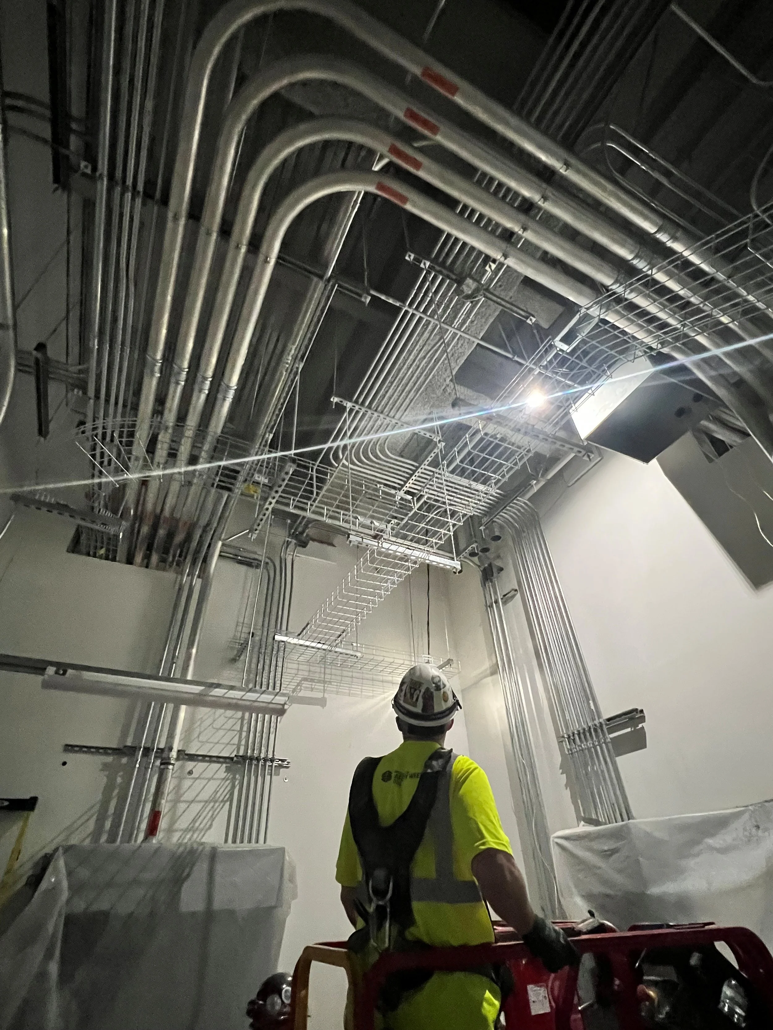 Construction worker in a hard hat and yellow safety vest standing in front of a complex network of electrical conduits and wiring in an indoor setting.