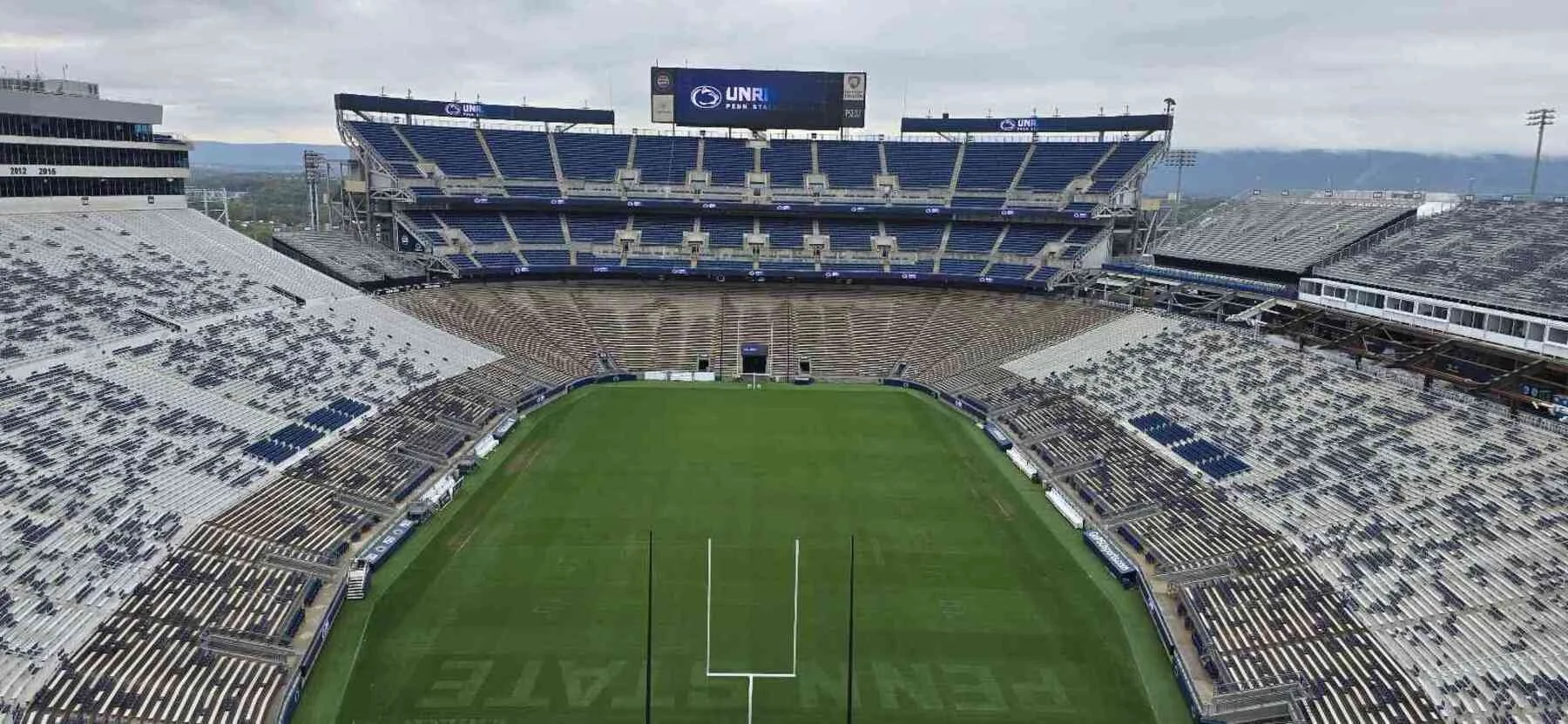 Empty football stadium with green field and tall empty stands, under an overcast sky, with a scoreboard displaying 'UNR' and 'Penn State' logos.