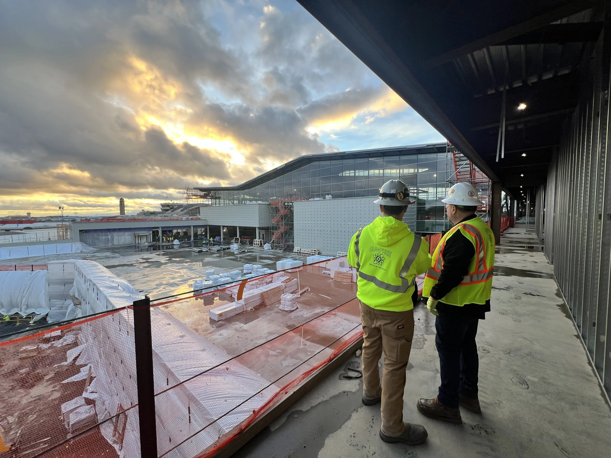 Two construction workers wearing hard hats and safety vests stand on a balcony viewing a construction site of a modern airport terminal at sunset.