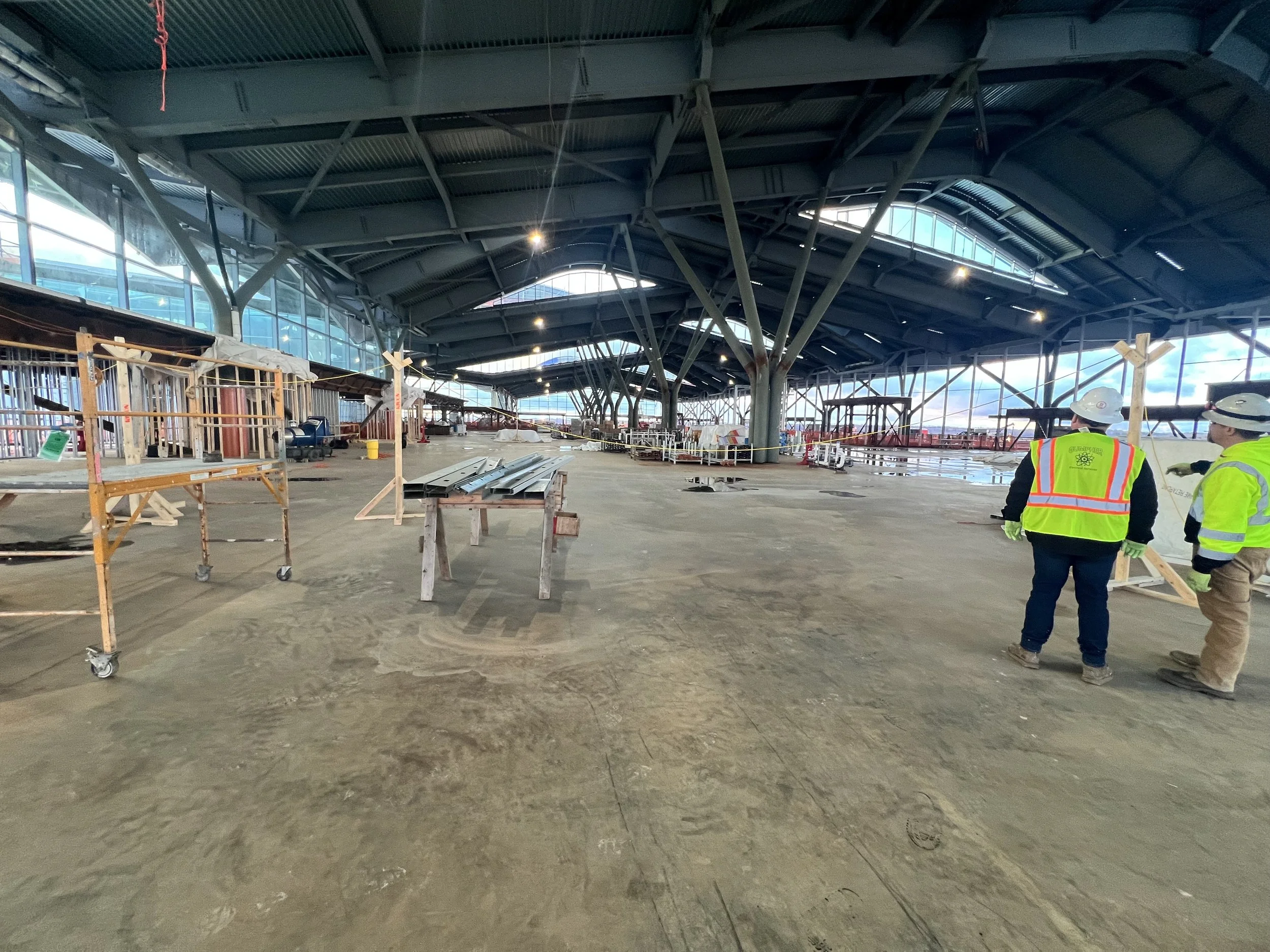 Interior view of a large construction site at an airport, with steel framework and glass windows. Two construction workers with helmets and safety vests are standing and discussing.