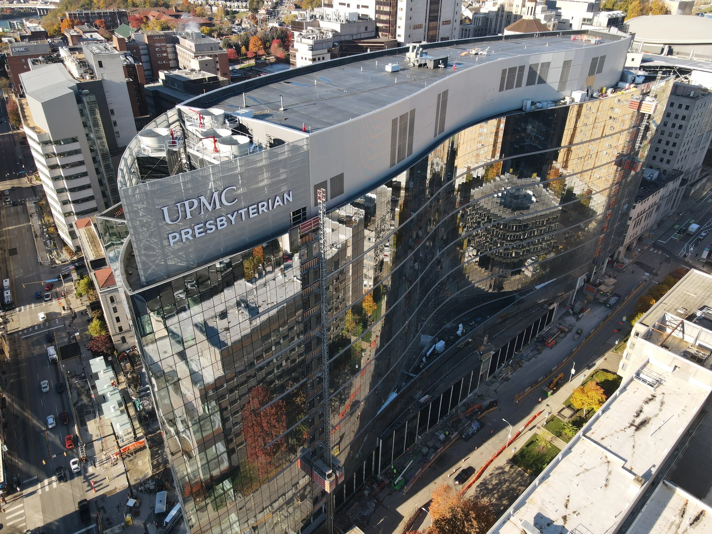 Aerial view of UPMC Presbyterian hospital building with reflective glass exterior, located in an urban area with streets, parked cars, and surrounding buildings.