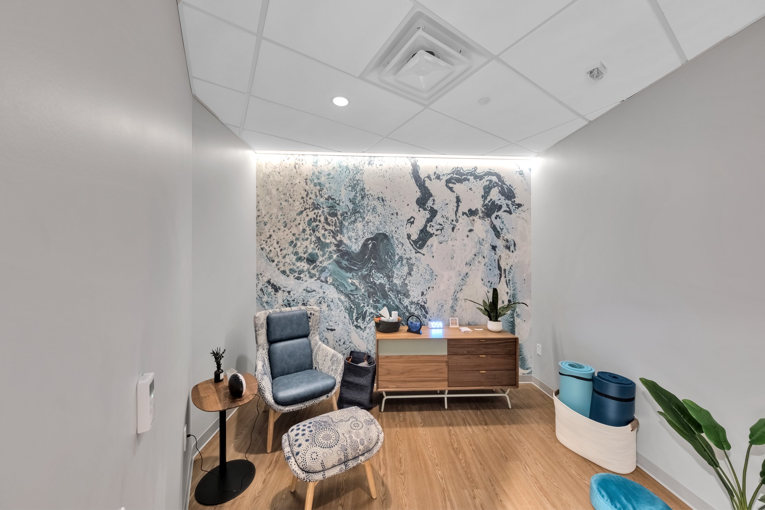 Modern office or waiting room with abstract wall art, a comfortable chair, a small side table with decorative items, a wooden cabinet with items and a plant, and exercise mats in a basket.
