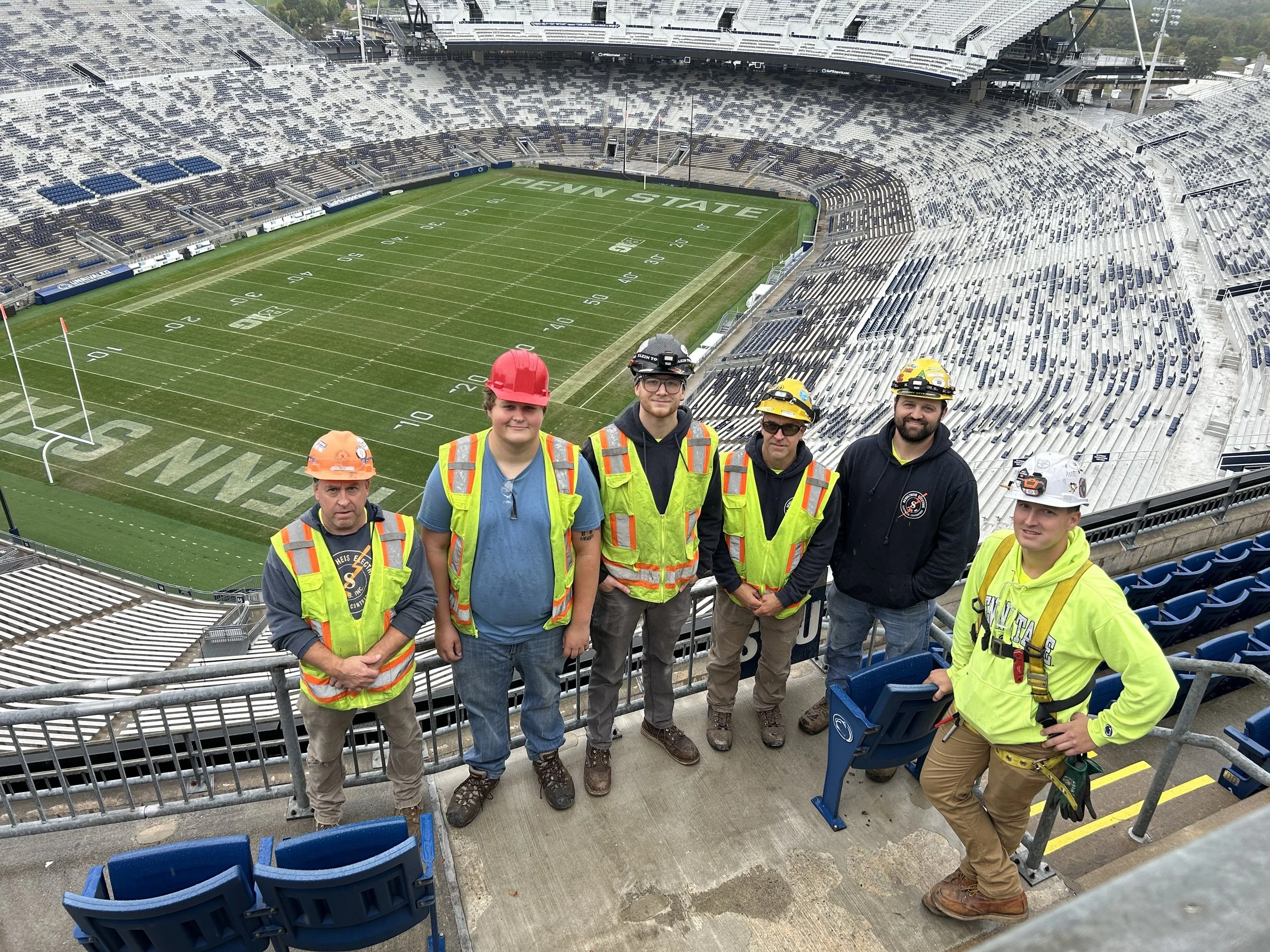 Six workers in safety gear stand on the bleachers at Penn State stadium, with the football field visible in the background.