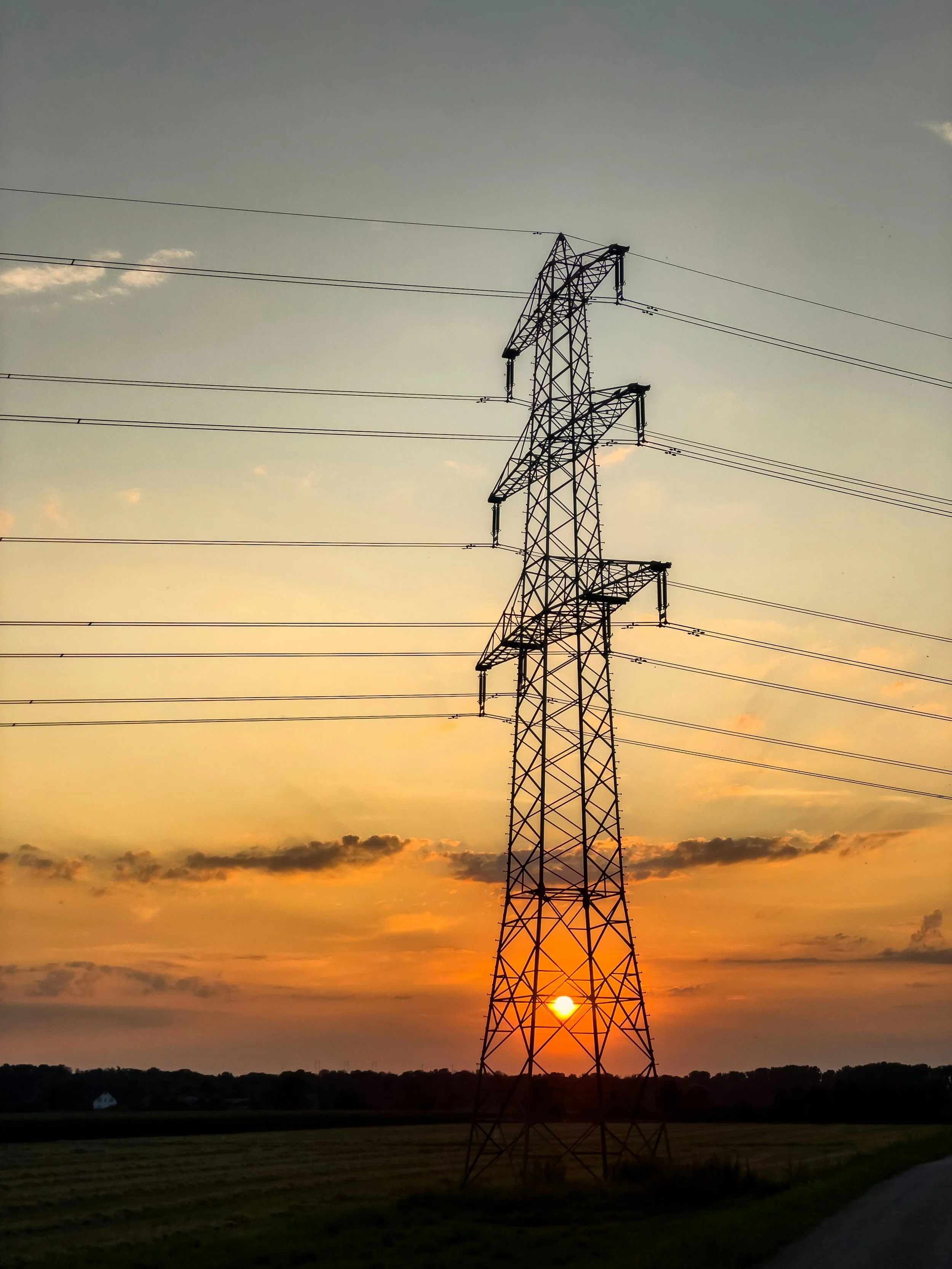 An electrical transmission tower standing in front of a sunset with the sun partially obscured by the tower, over a rural landscape with fields and trees.
