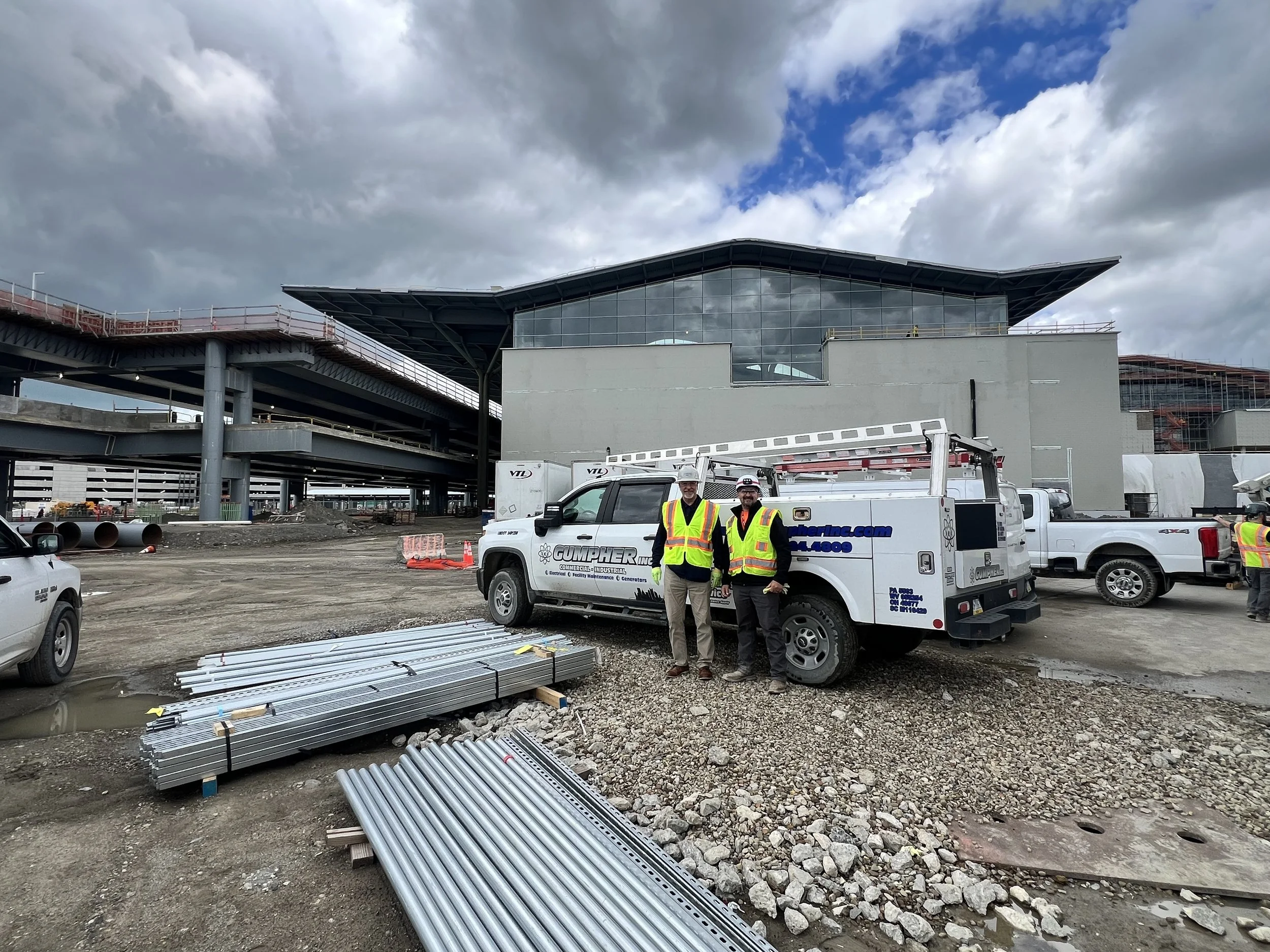Construction workers in high-visibility vests standing beside a work truck on a construction site with modern building and infrastructure in the background.