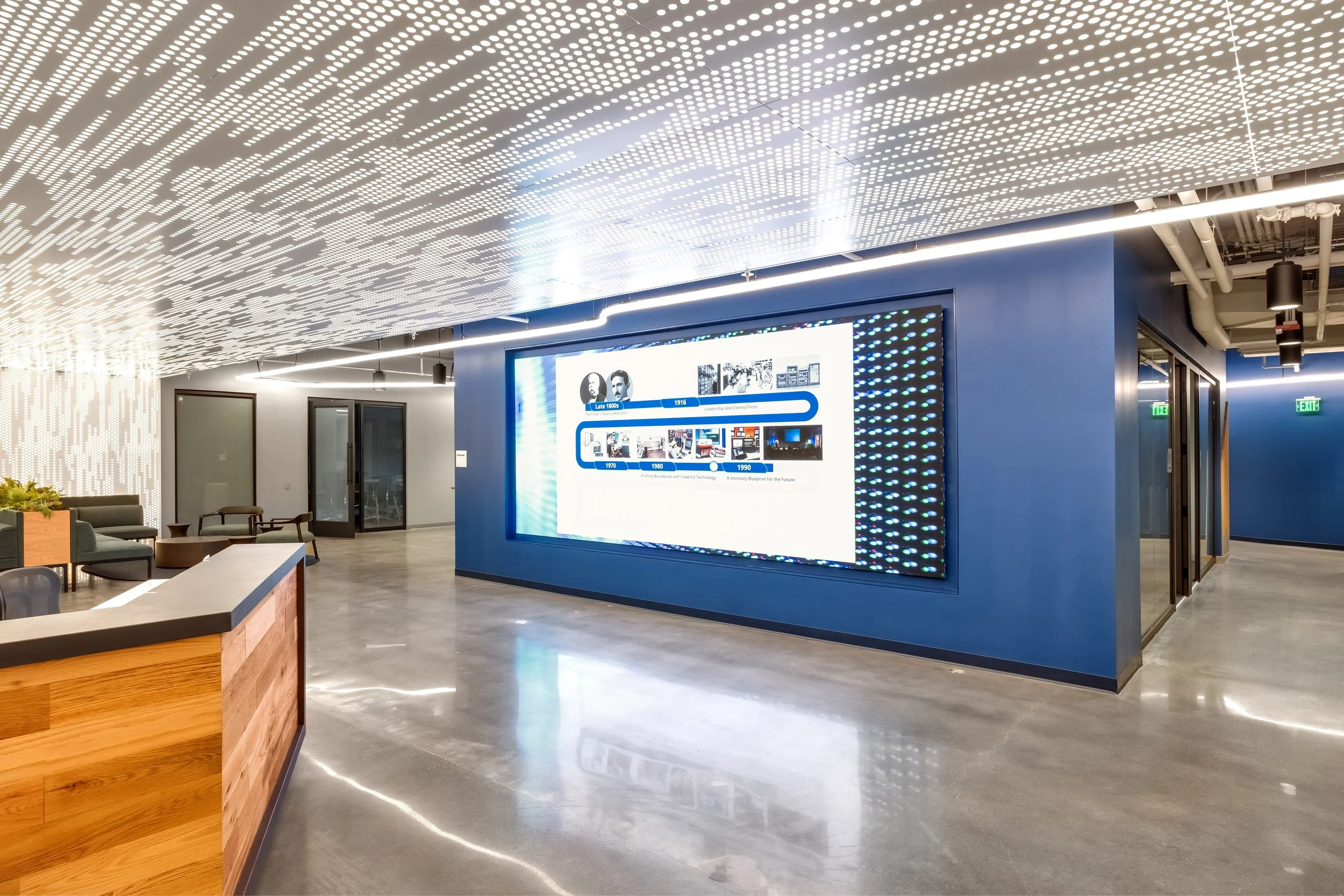 Modern office lobby with large digital display on blue wall, ceiling with artistic perforated design, polished concrete floor, seating area with chairs and tables, indoor plants, glass doors, and visible exit signs.