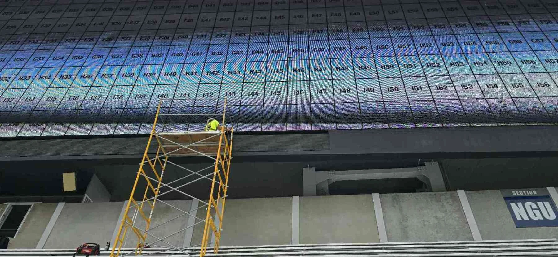 A worker in a safety vest standing on scaffolding, cleaning or working on a large digital billboard displaying a spreadsheet or table with rows and columns of data.