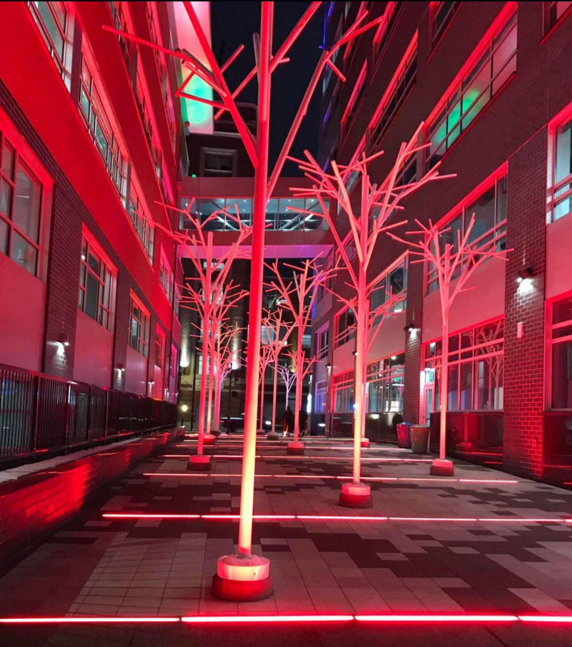 Nighttime view of an outdoor walkway illuminated by red lights on stylized leafless trees, with surrounding modern brick and glass buildings.