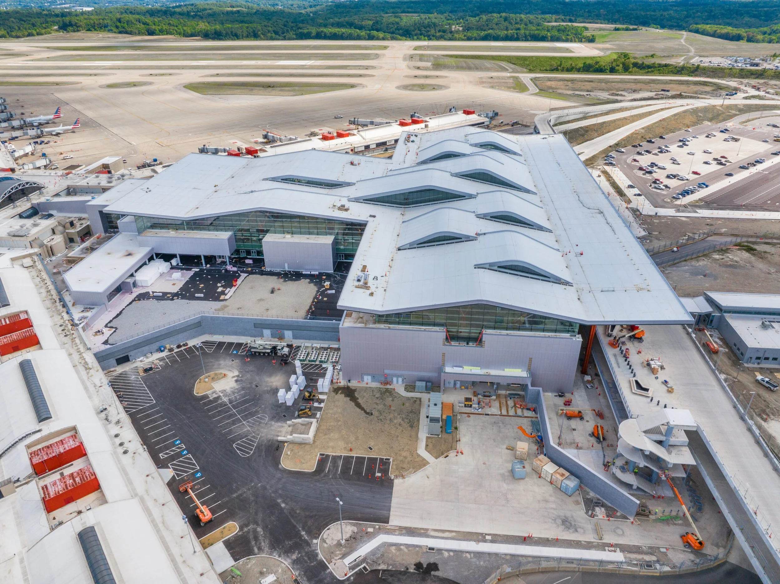 Aerial view of a modern airport terminal under construction, with empty parking lots, construction equipment, and airplanes parked at gates nearby.