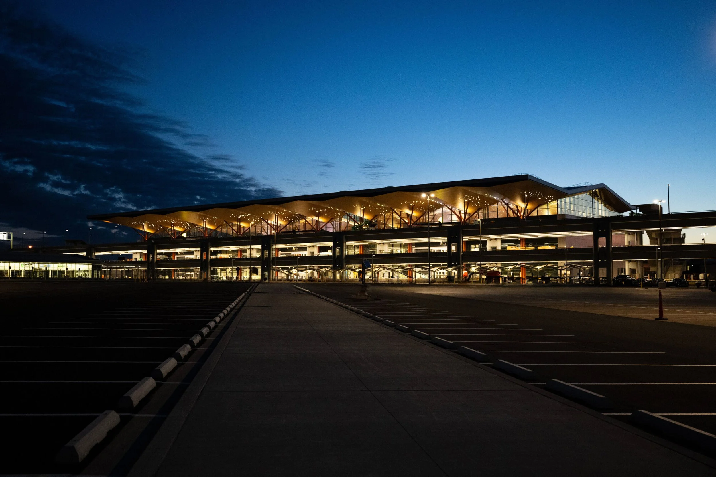 An illuminated modern airport terminal building at dusk with a large parking lot in the foreground and a darkening sky in the background.