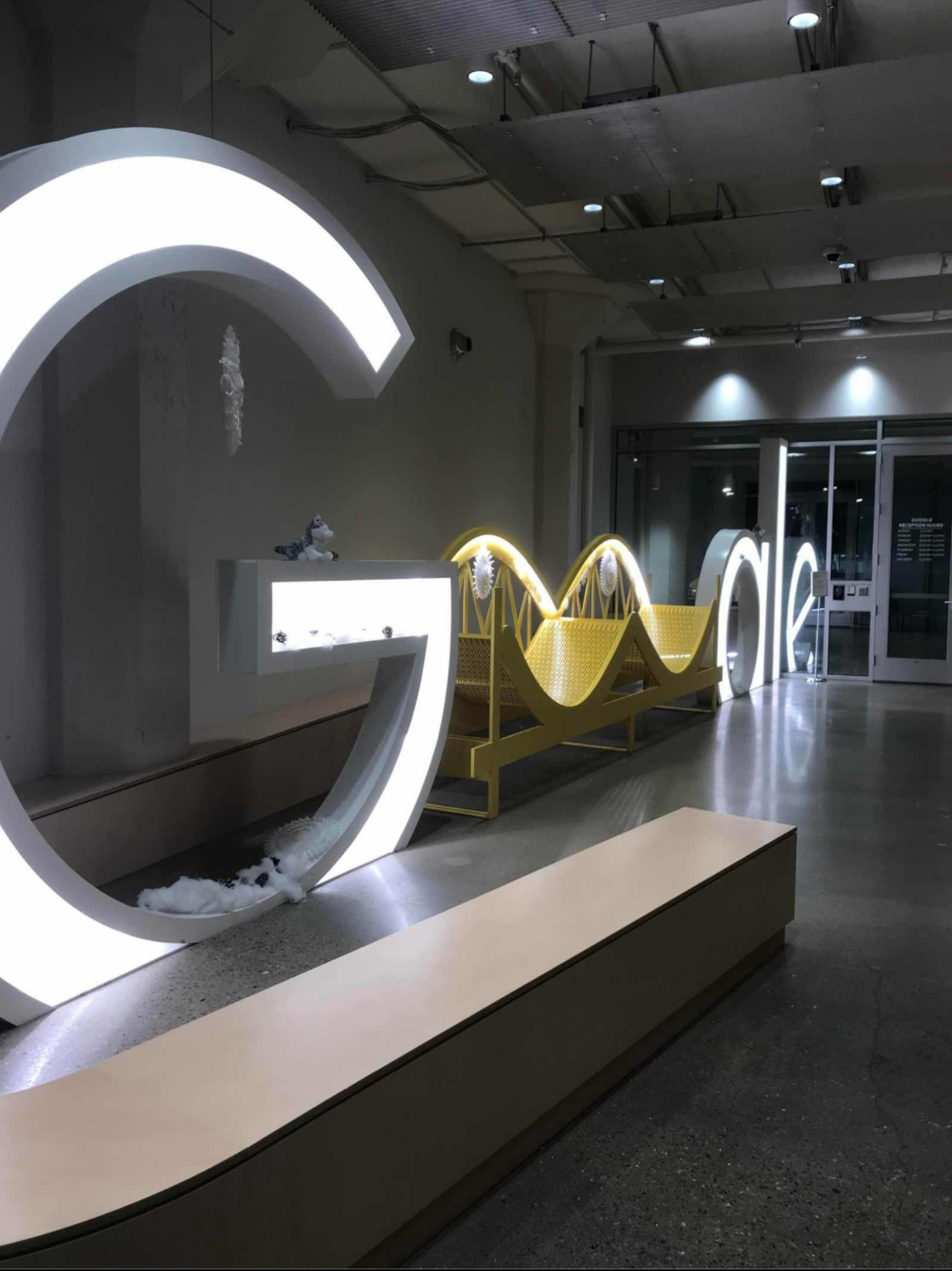 Illuminated letters spelling 'GLOW' with a yellow and white color scheme, displayed inside a modern building with a polished concrete floor and an open entrance in the background.