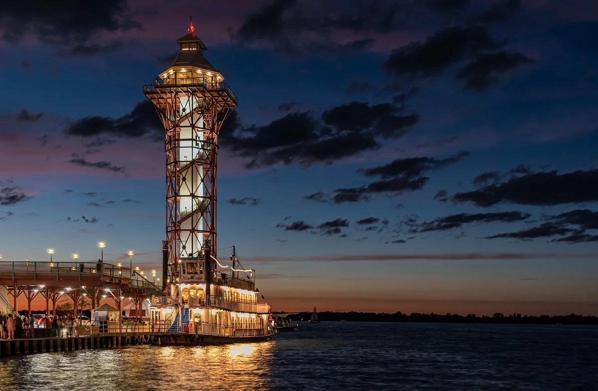 Lighthouse tower along a river at sunset with boats passing by