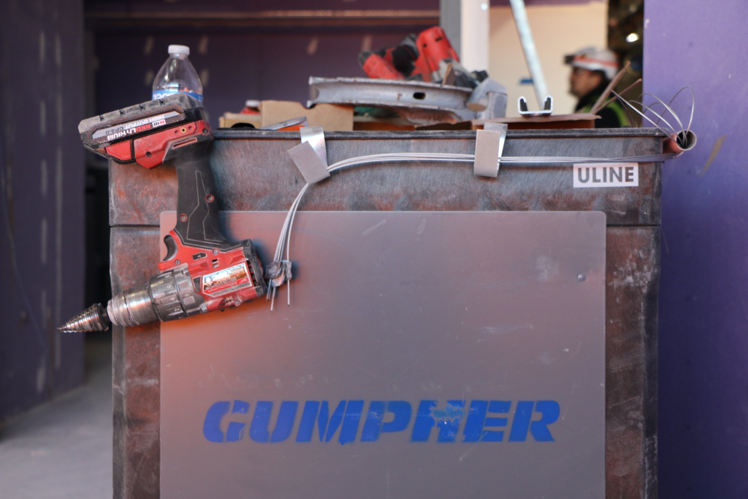 Construction tool box with a cordless drill, wires, and other tools on top, with a construction worker in safety gear in the background.