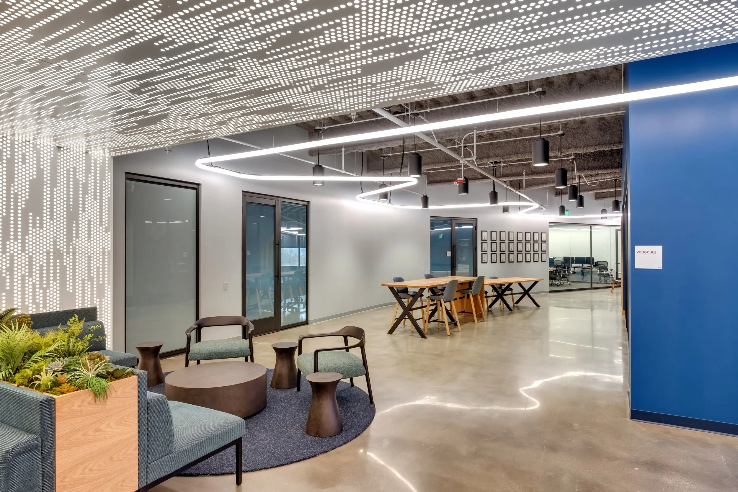 Modern office lobby with seating area, plants, and a visitor hub sign, featuring a polished concrete floor, white walls, large glass doors, and contemporary lighting fixtures.