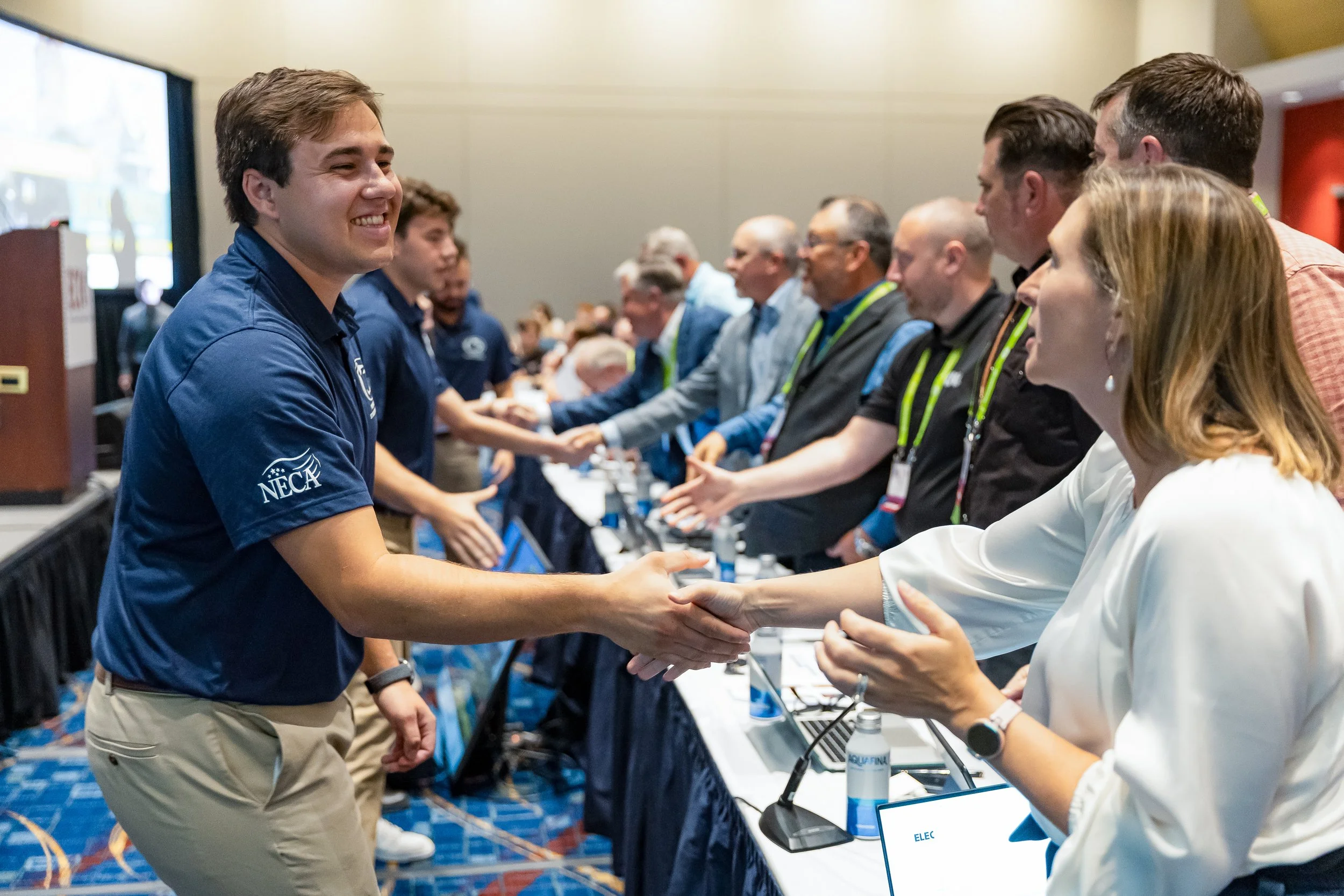 Young man in a blue shirt shaking hands with an older woman at an academic conference, with other people in line shaking hands in the background.