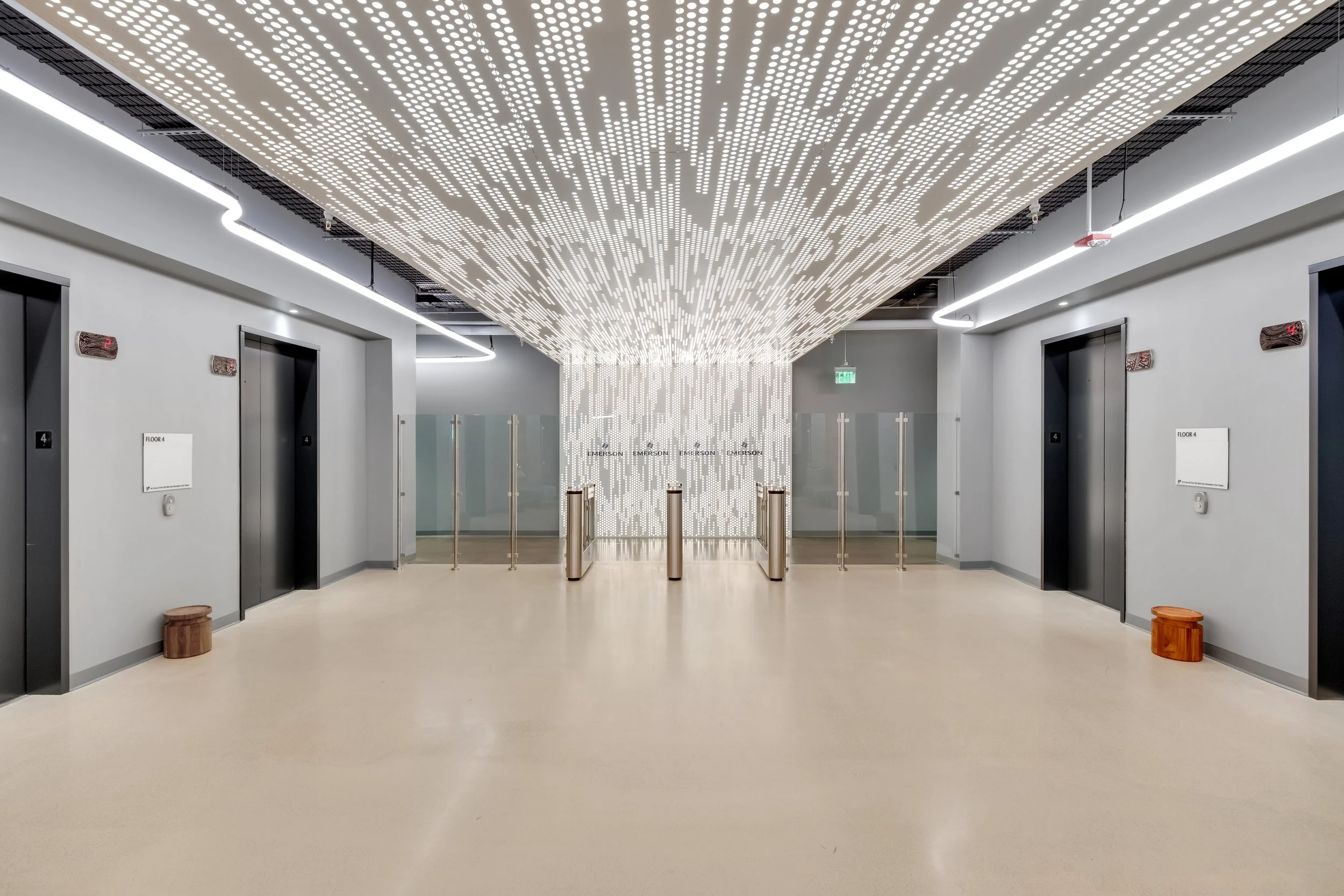 Modern elevator lobby with four elevators, illuminated ceiling installation, glass doors at the rear, and minimalistic decor.