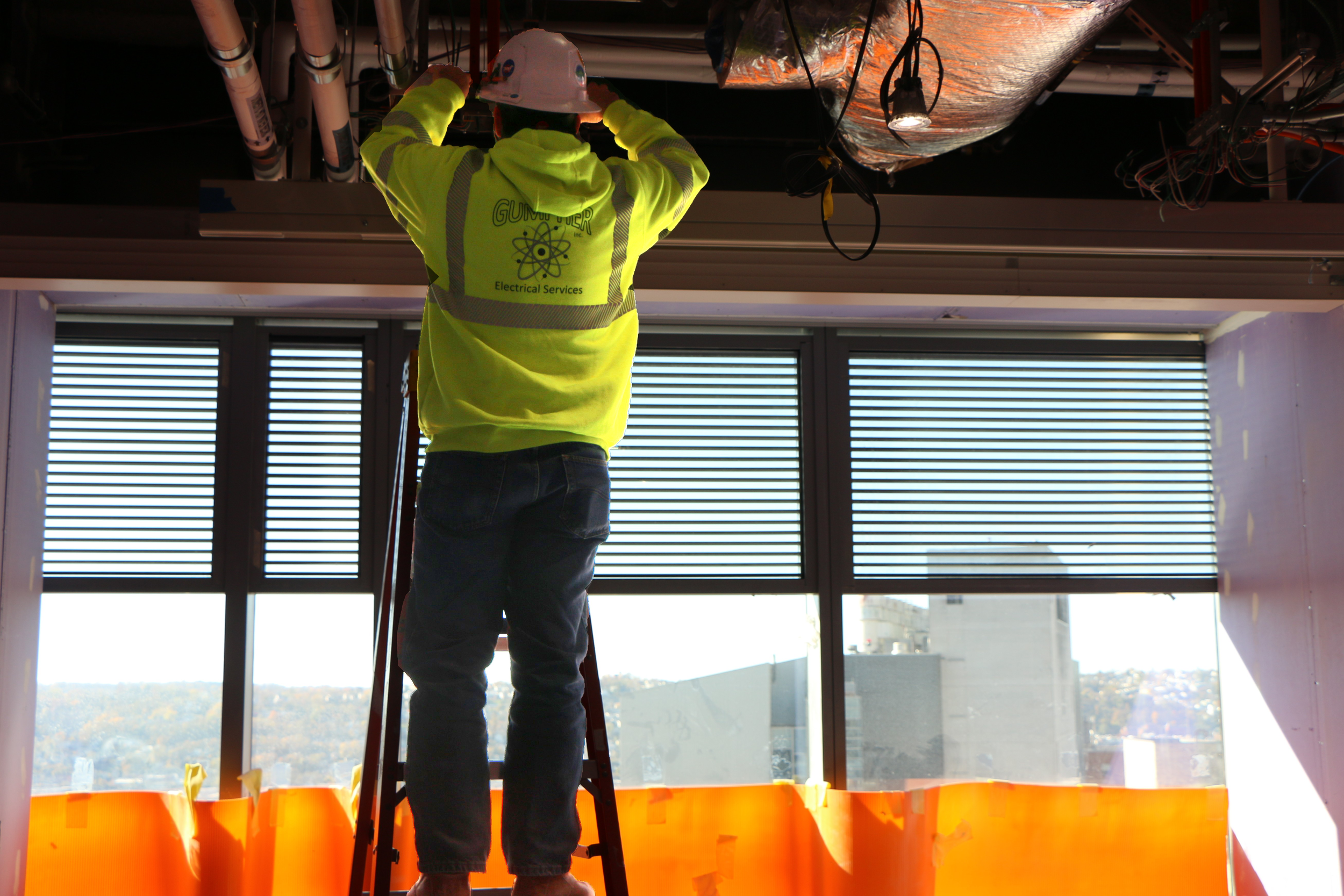 Electrician in a bright yellow safety jacket and white helmet working on electrical wiring on the ceiling of a building with large windows showing a cityscape, using a ladder.