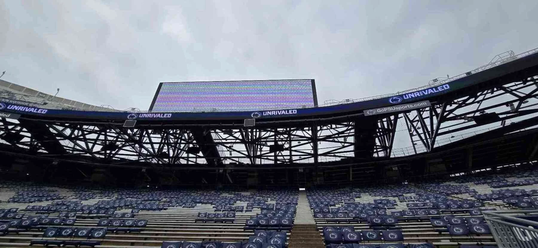 Interior view of a sports stadium with empty seating, a large digital scoreboard, and electronic advertising banners for UNRIVALED and GoPSUsports.com