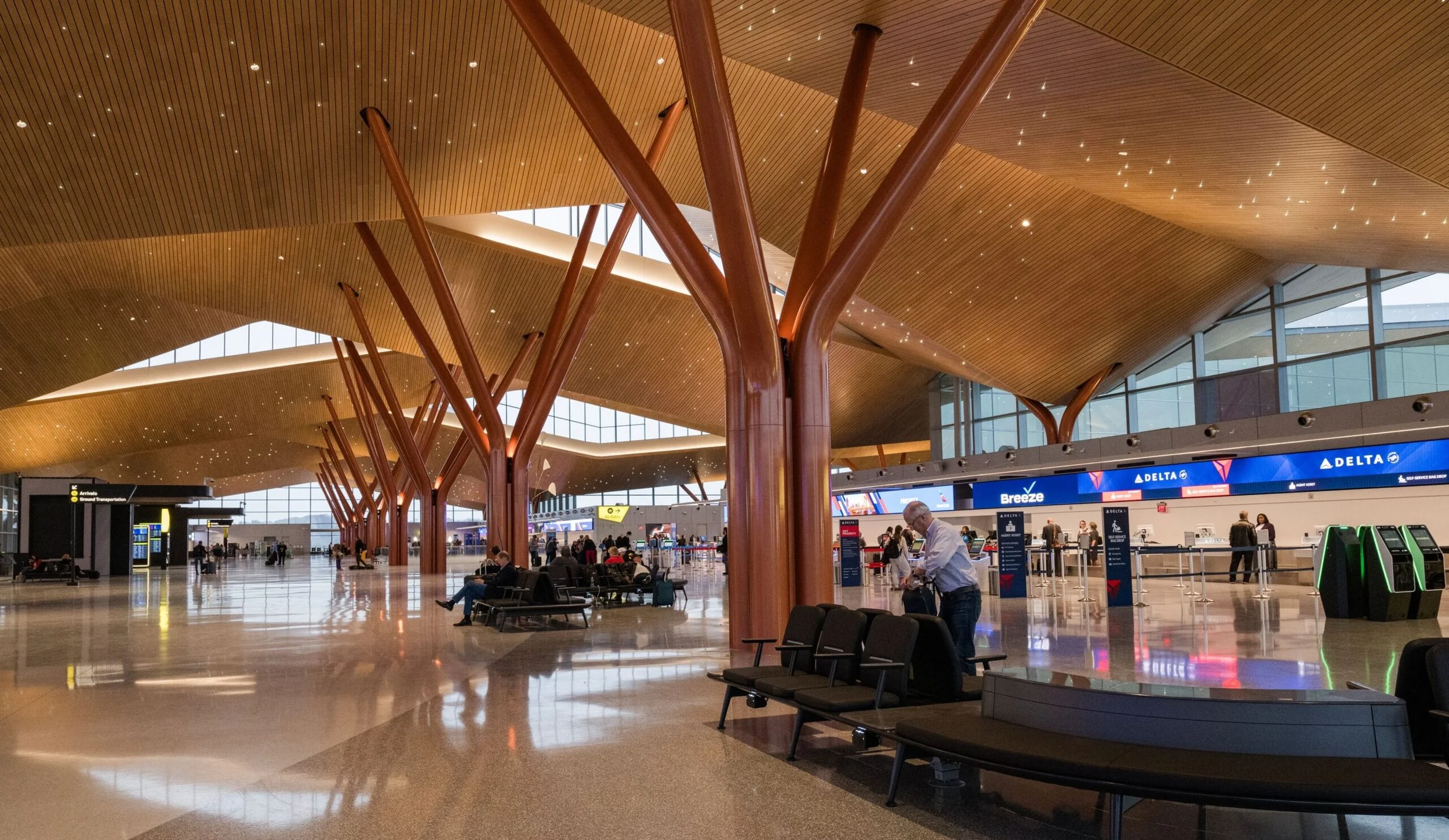 Interior of an airport terminal with wooden tree-like columns, high ceilings, and large windows. Passengers are seated and walking, with a Delta Airlines check-in area visible in the background.